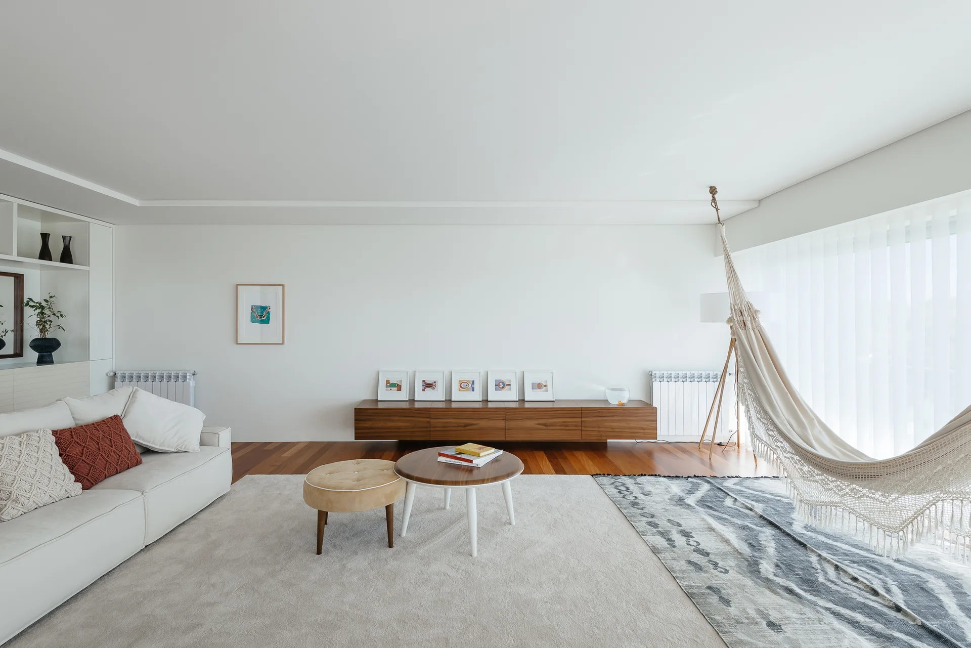 A minimalist white living room features a white sofa, a low wood console with framed art, and a woven fringe hammock near sheer curtains.