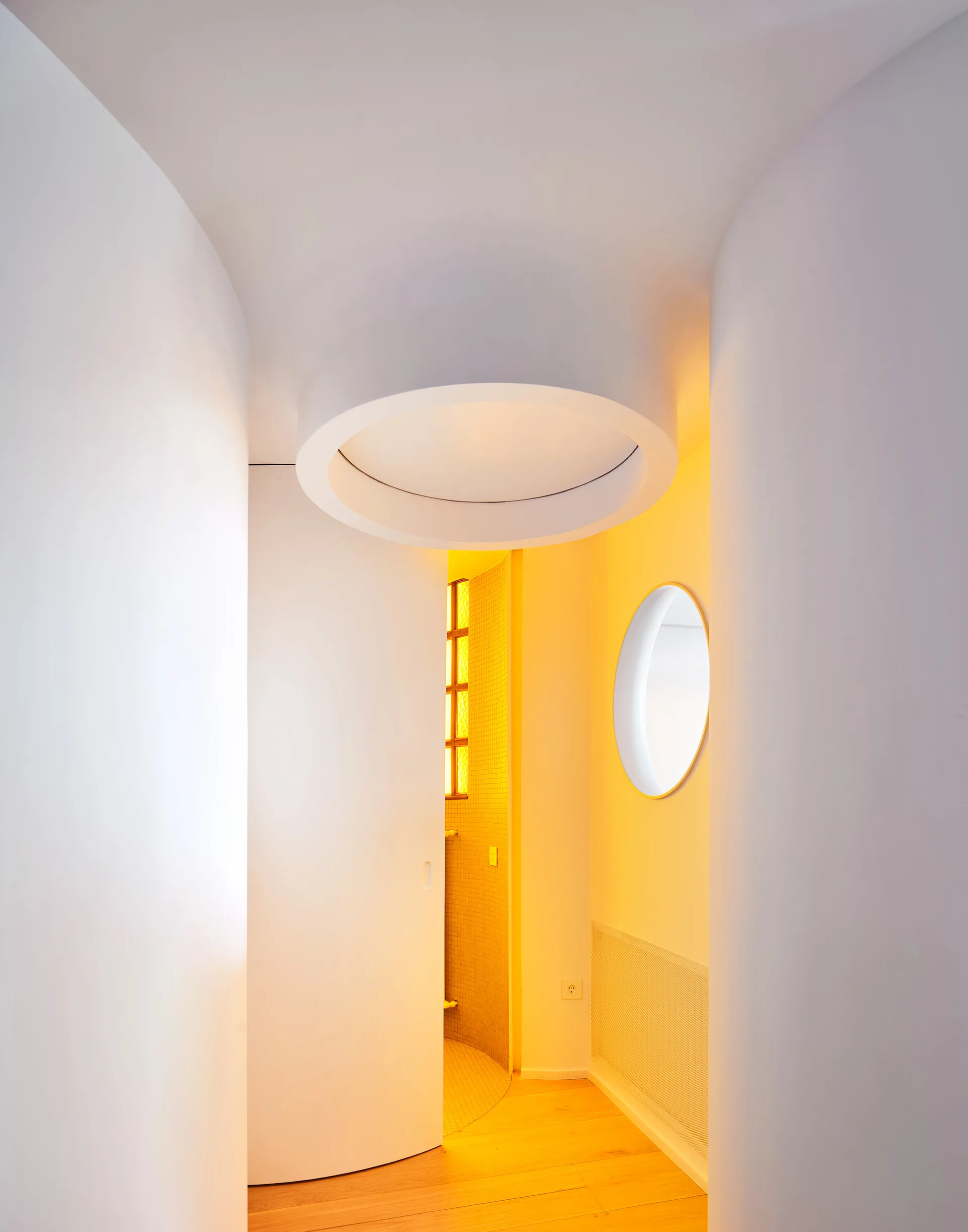 A minimalist white hallway featuring curved walls, a circular recessed ceiling light, a round wall mirror, and warm yellow light glowing from an adjacent room.