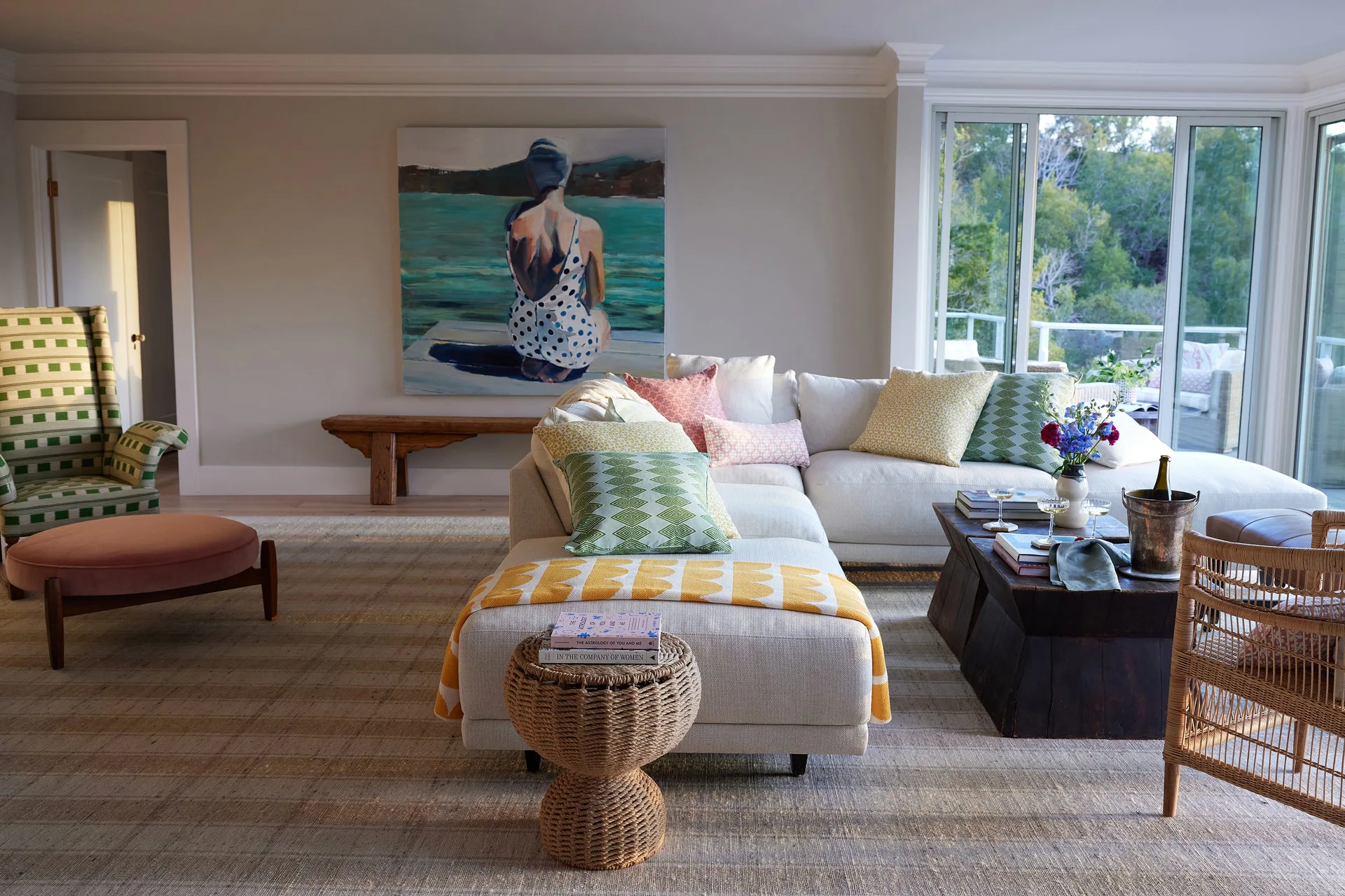 A bright living room features a cream sectional sofa with colorful pillows, a checkered armchair, and a large painting of a swimmer.