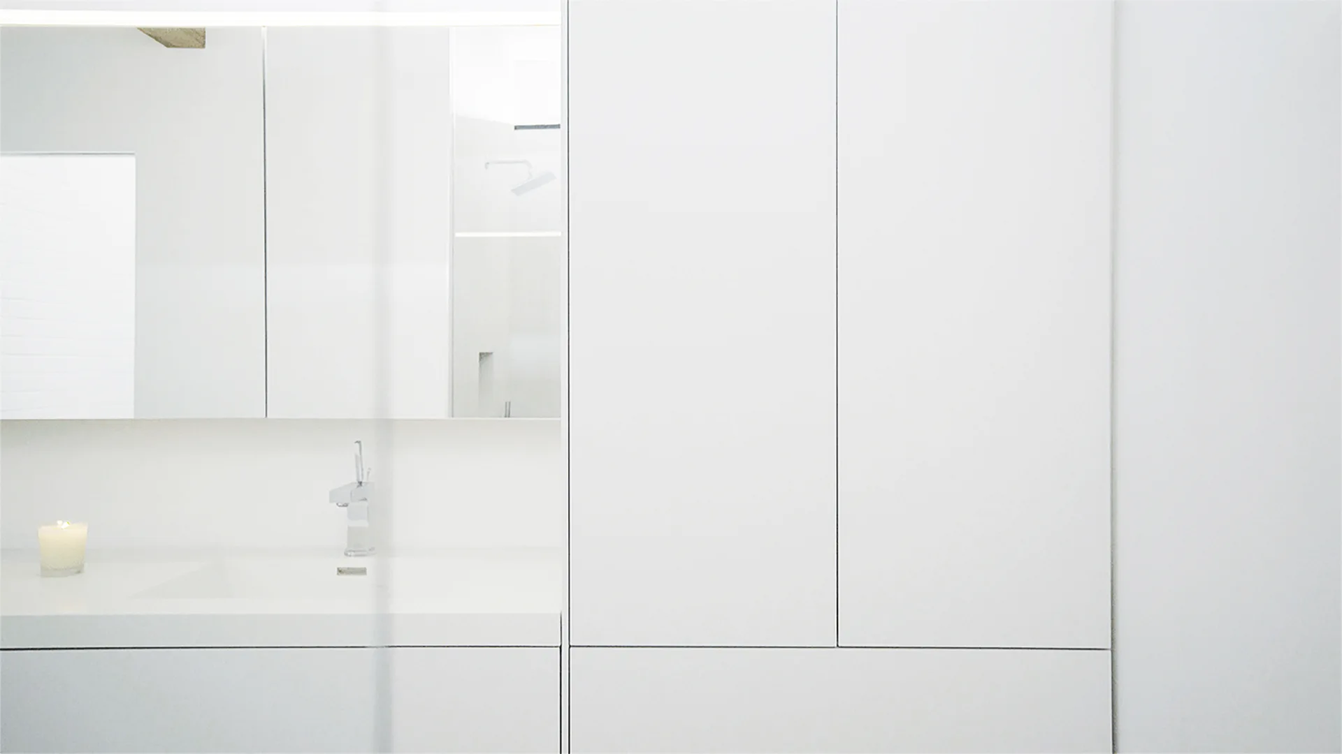 A minimalist white bathroom features an integrated sink vanity, a large frameless mirror, and seamless full-height storage cabinets.