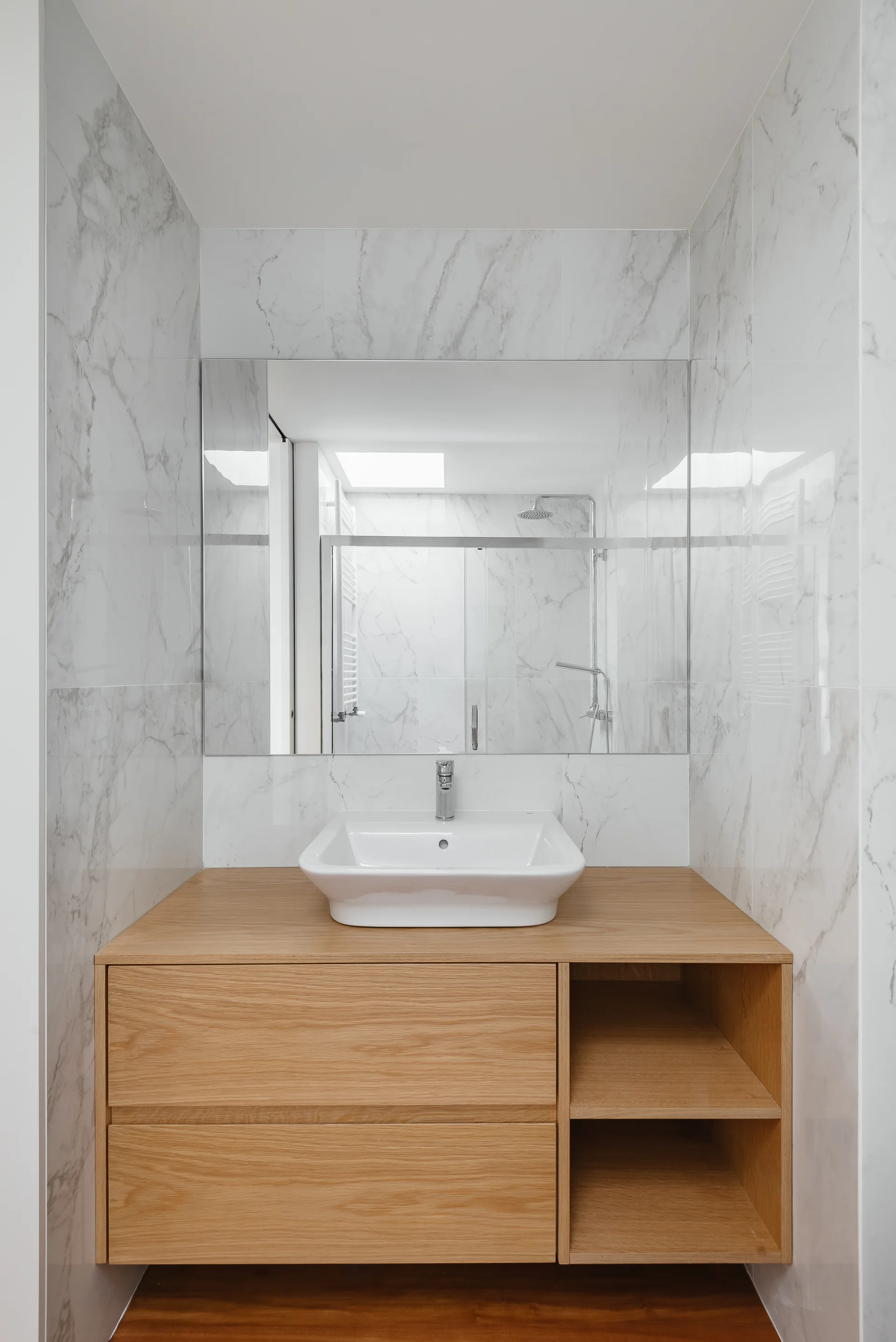 A floating light wood bathroom vanity with a white ceramic sink is set against veined white marble wall tiles beneath a large frameless mirror.