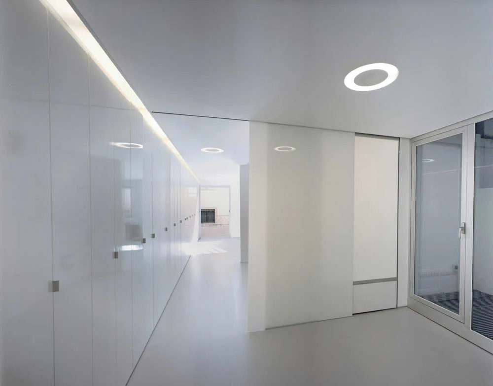 A bright white minimalist hallway features a long wall of glossy handleless cabinets, recessed ceiling downlights, and a glass door.