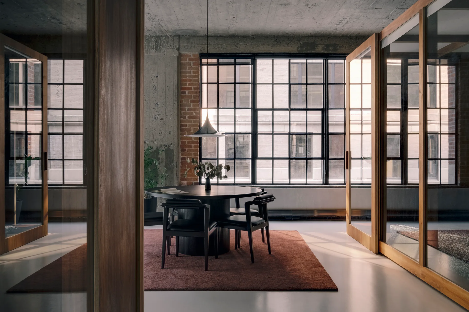 A black dining set sits on a rust rug in an industrial loft with concrete ceilings, a large grid window, and wood-framed glass partitions.