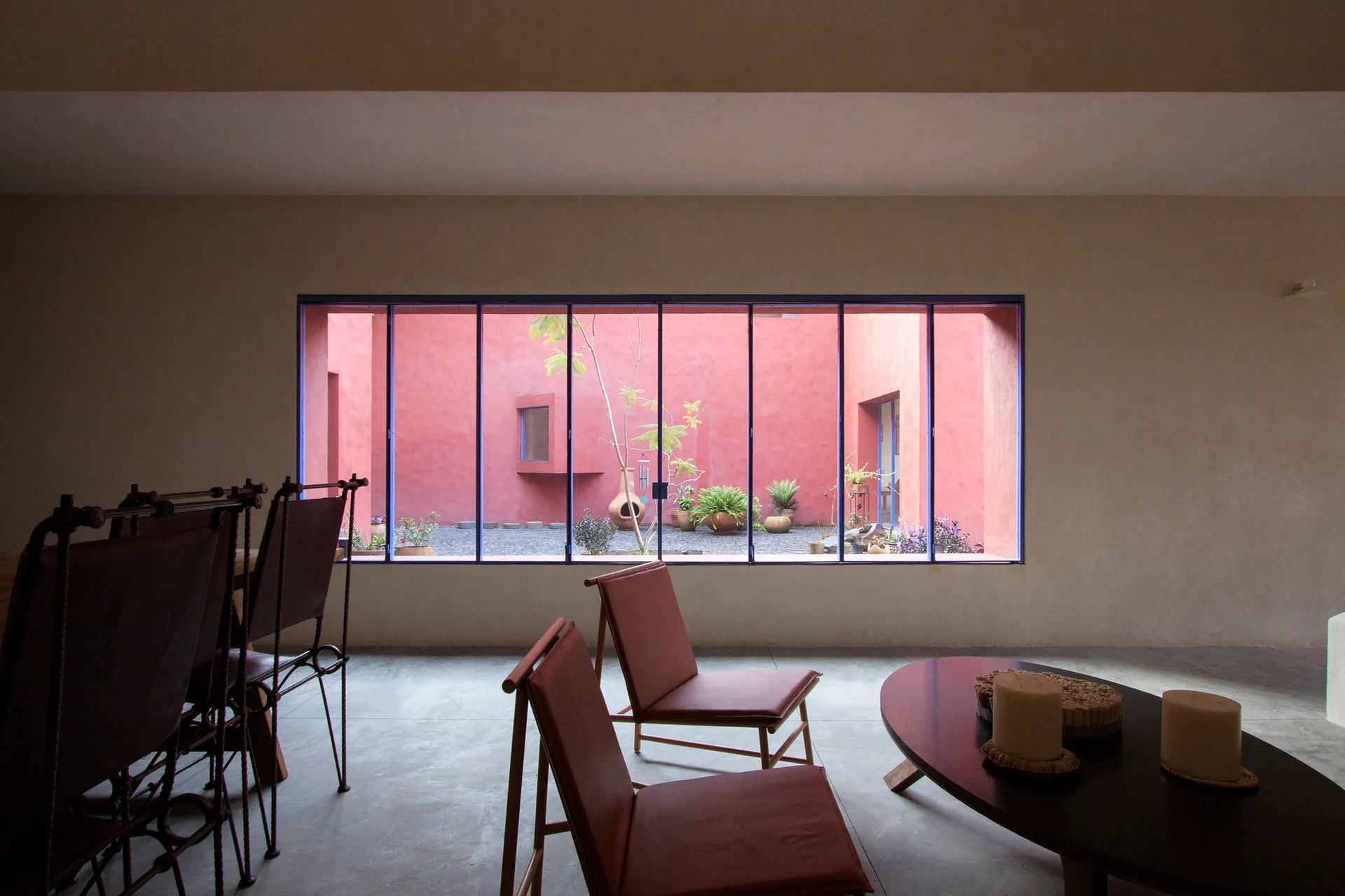 A minimalist living room with low brown leather chairs and a dark wood coffee table facing a large window that frames a red-walled outdoor courtyard.