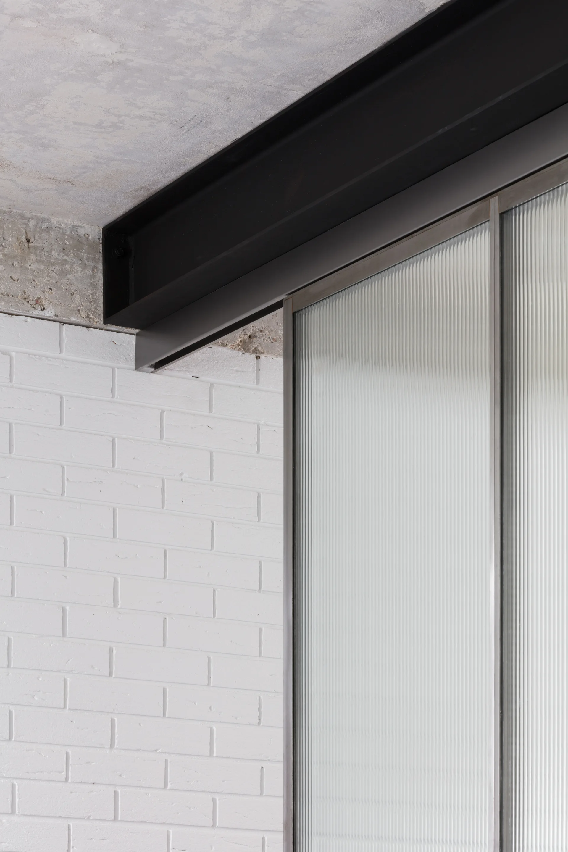 Architectural detail featuring a black steel ceiling beam intersecting a white painted brick wall next to a textured reeded glass partition panel.