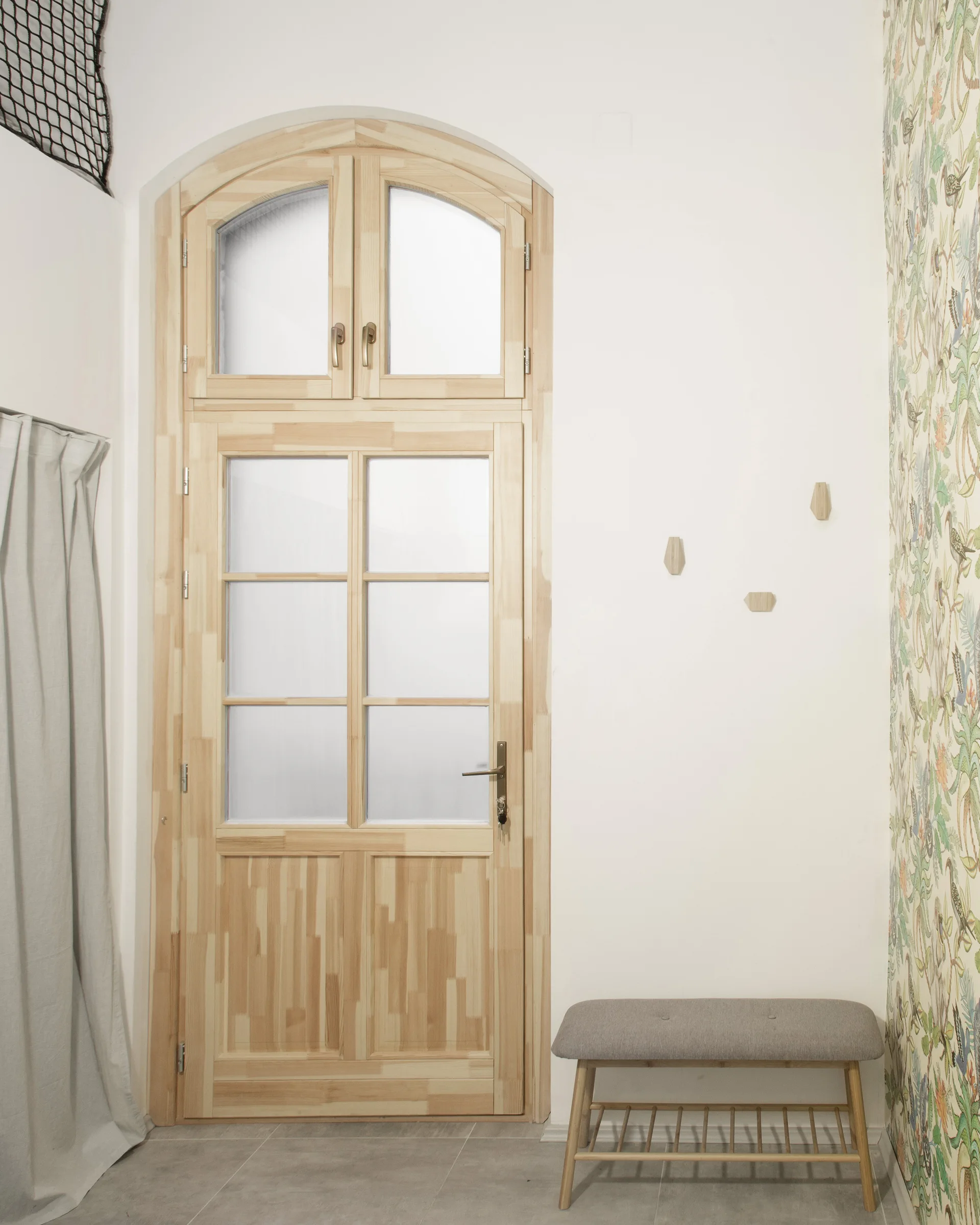 An arched light wood door with glass panes stands next to a grey cushioned bench and botanical print wallpaper.