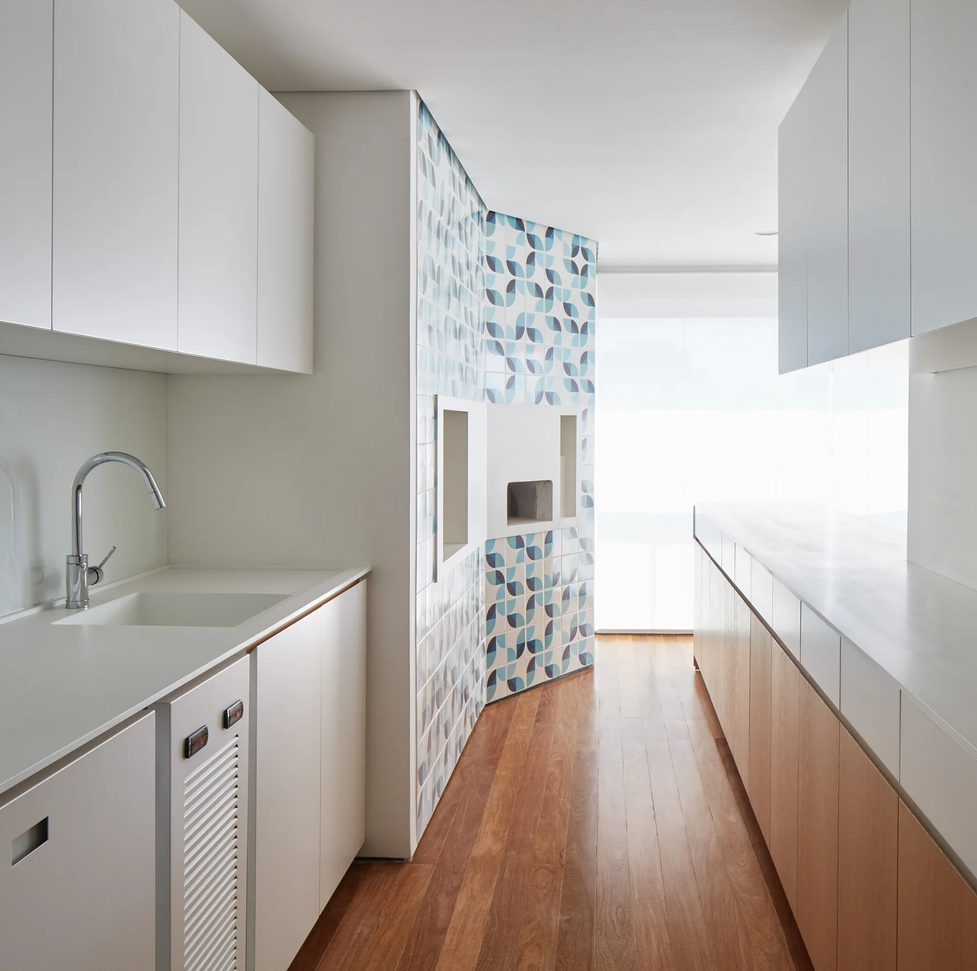 A galley kitchen featuring white upper cabinets, wood base cabinets, a medium-toned wood floor, and a prominent blue and white geometric tiled wall.