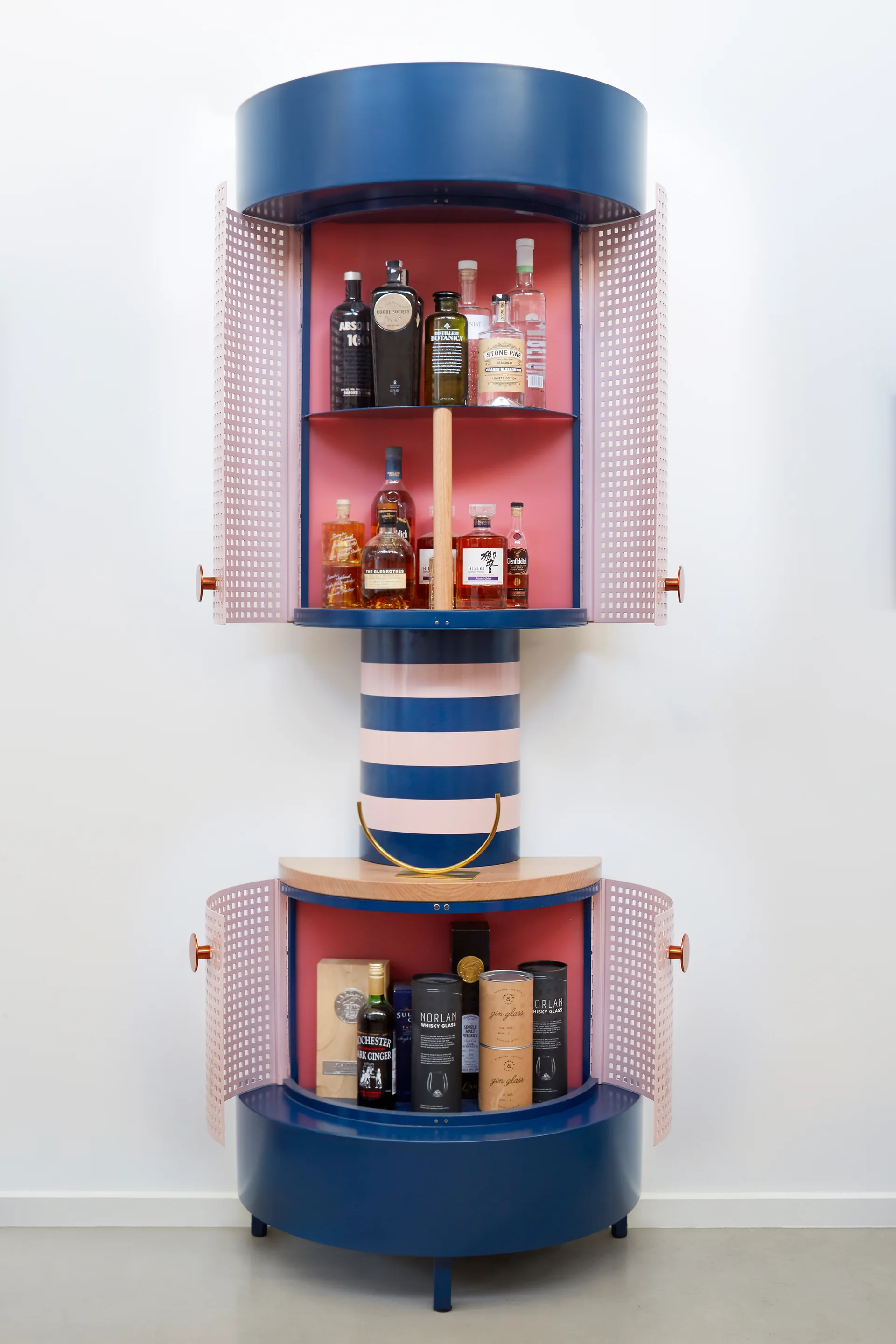 A cylindrical two-tier bar cabinet featuring a navy blue frame, light pink perforated metal doors, and striped central column holds liquor bottles.