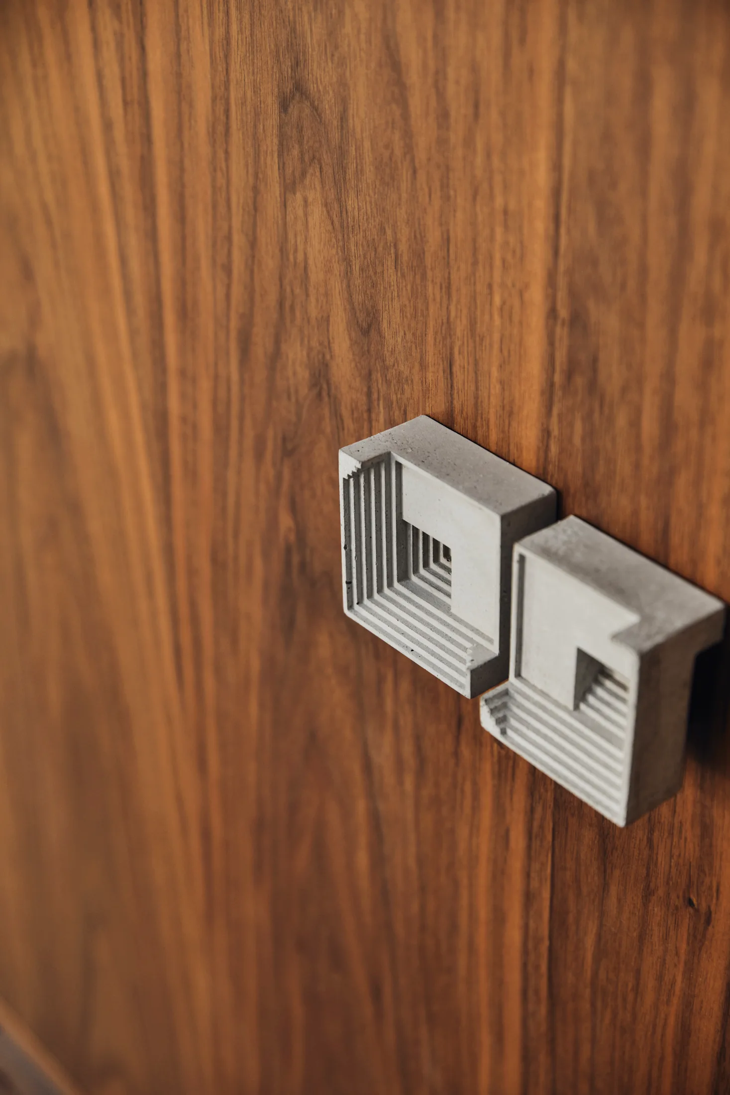 Two grey concrete drawer pulls featuring a stepped geometric design are mounted on a smooth brown wood cabinet door.