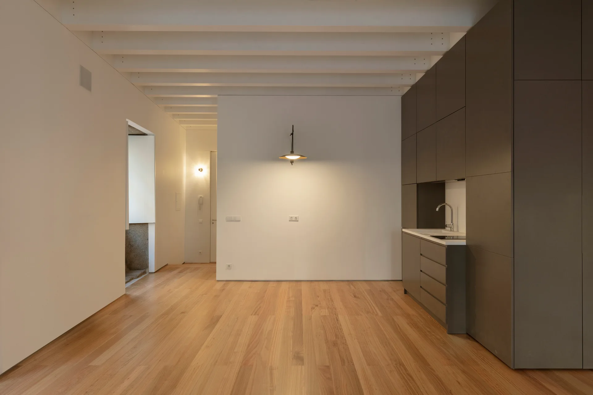 A minimalist studio interior showing a dark grey integrated kitchenette set against plain white walls with light wood flooring and exposed ceiling beams.