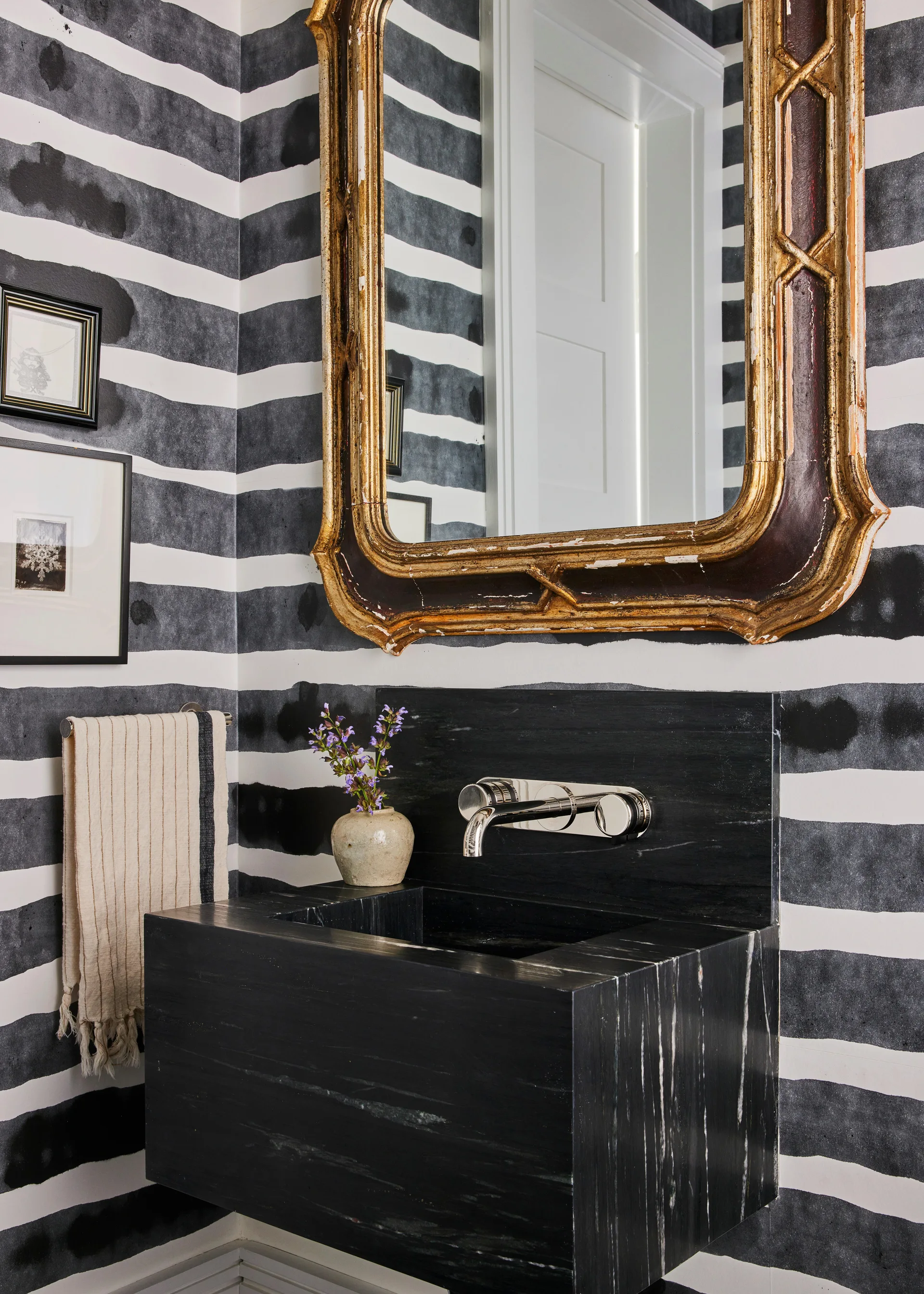 A modern powder room features a wall-mounted black stone block sink beneath a gilded mirror on horizontally striped wallpaper.