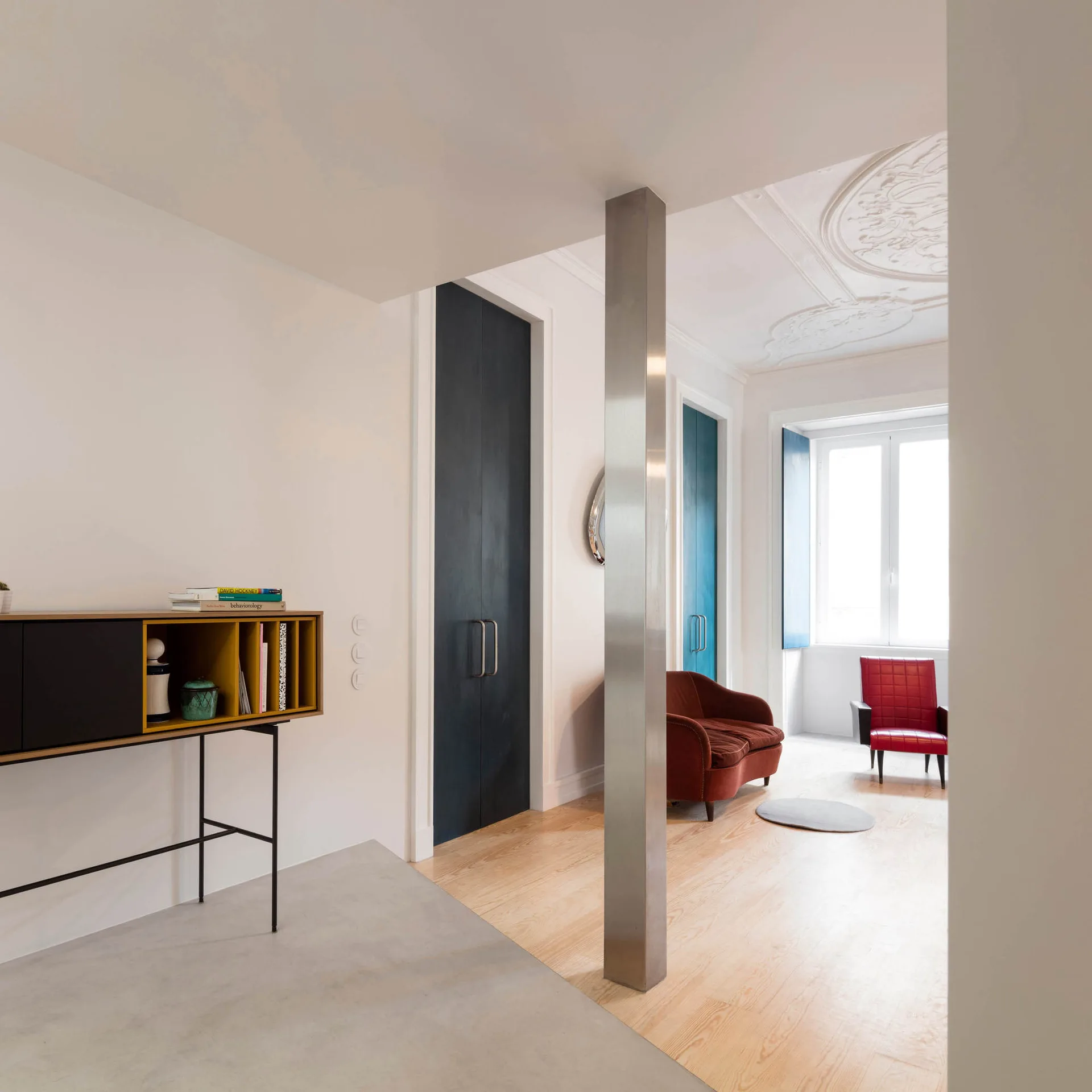 A bright living space with light wood flooring, a central square steel column, tall dark blue doors, and a decorative plaster ceiling medallion.