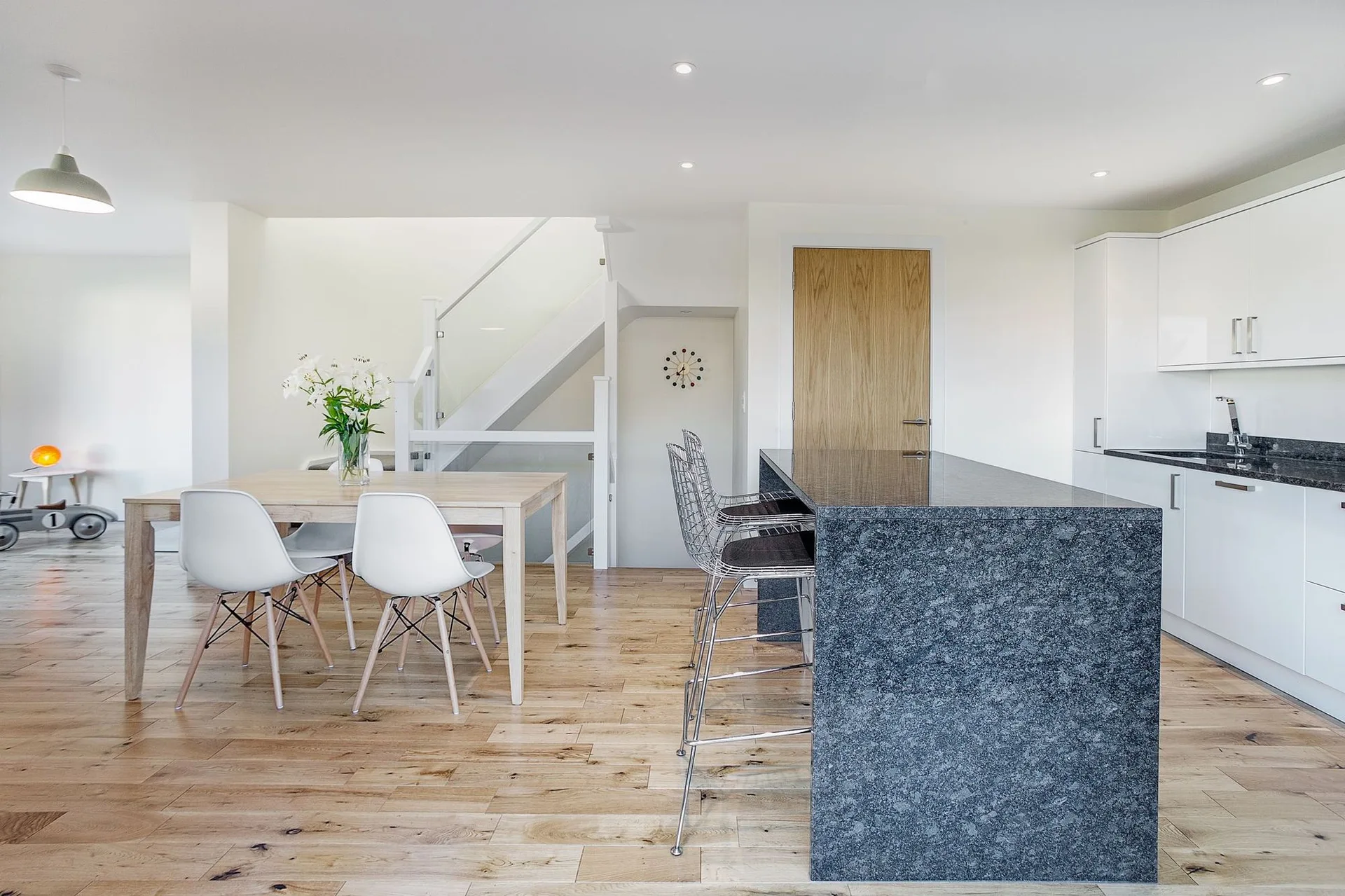 A modern open-plan kitchen and dining area with a grey stone island, metal wire stools, a wood dining table, and a glass-railing staircase.