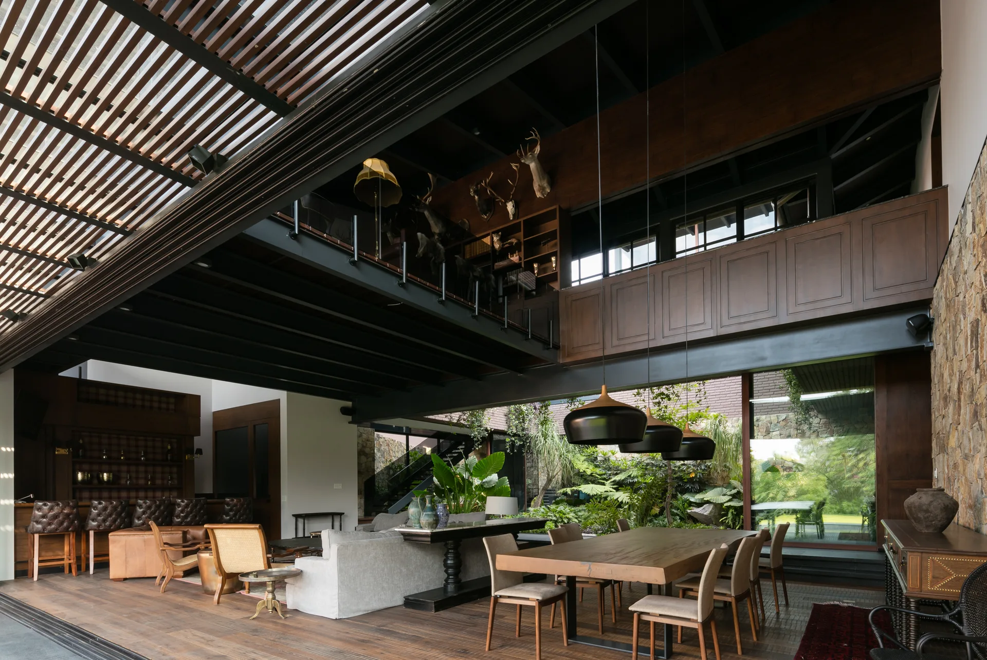 A spacious open-plan living and dining area with a wood table, grey sofa, stone wall, and a mezzanine level overlooking an indoor-outdoor garden.