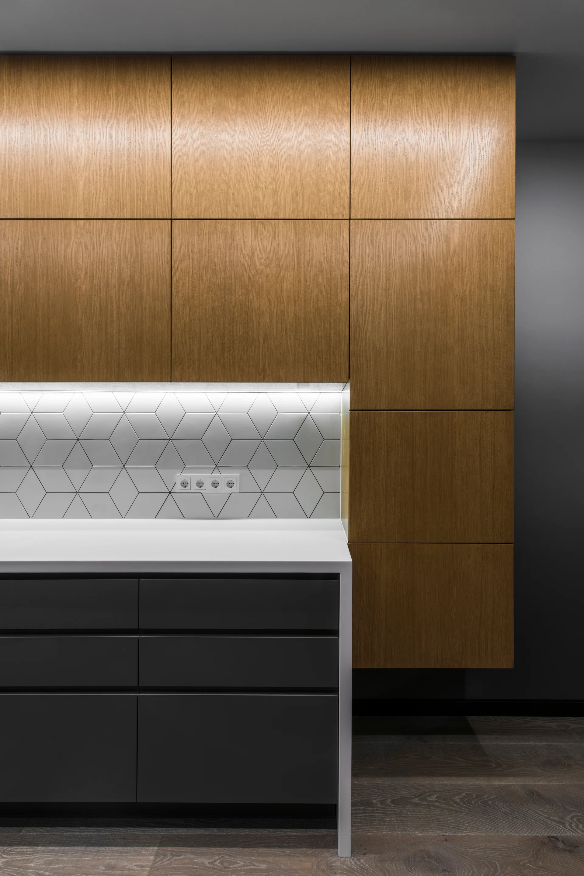 Modern kitchen featuring warm oak upper cabinets, charcoal gray lower drawers, a white countertop, and a geometric tile backsplash with integrated lighting.