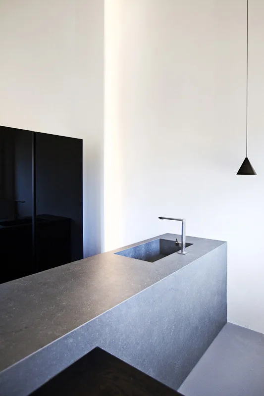 A minimalist kitchen features a monolithic grey stone island with an integrated sink, a single black cone pendant light, and a vertical warm light niche.