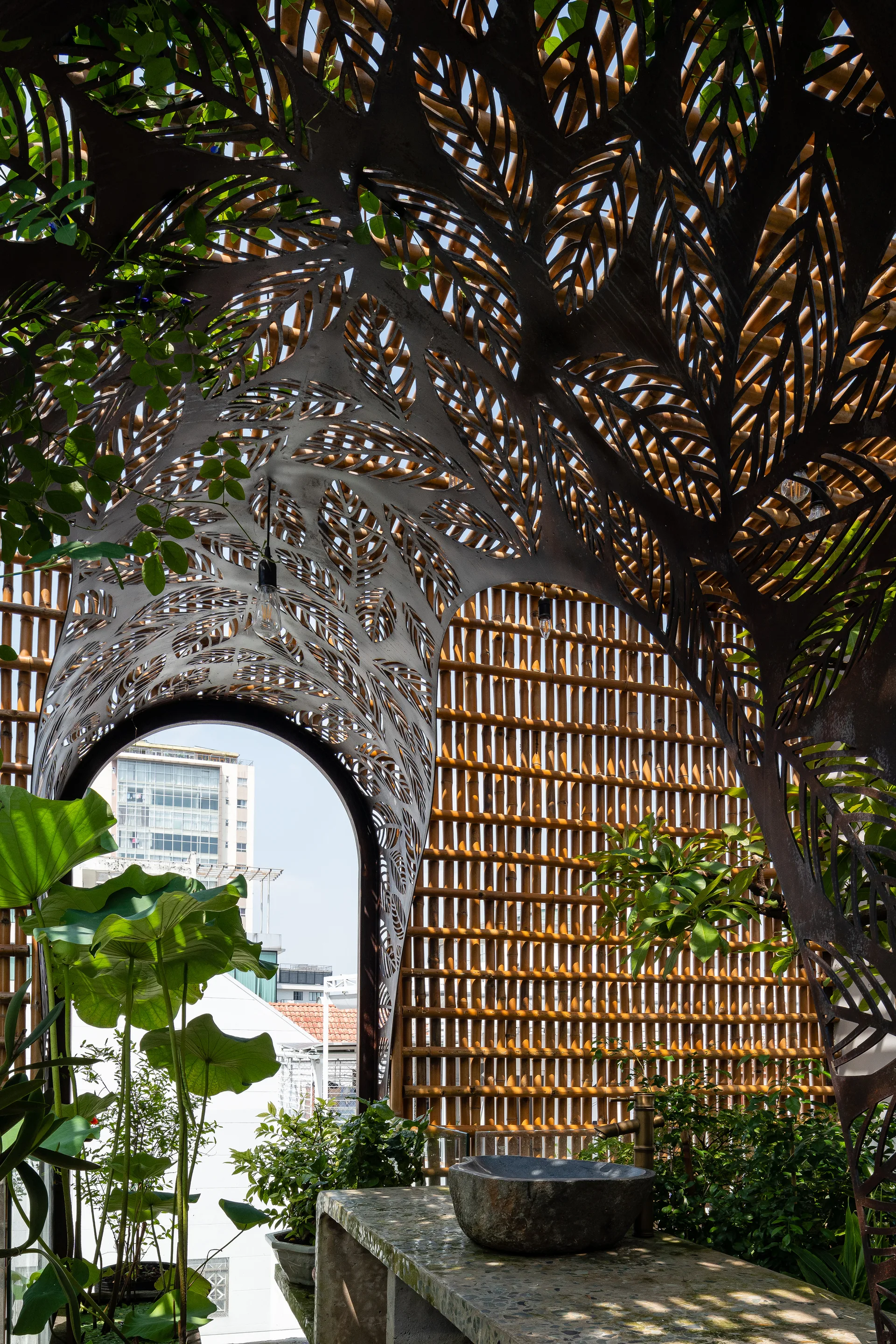 An outdoor patio features a stone basin sink set against a vertical bamboo rod wall under an intricate leaf-patterned metal canopy.