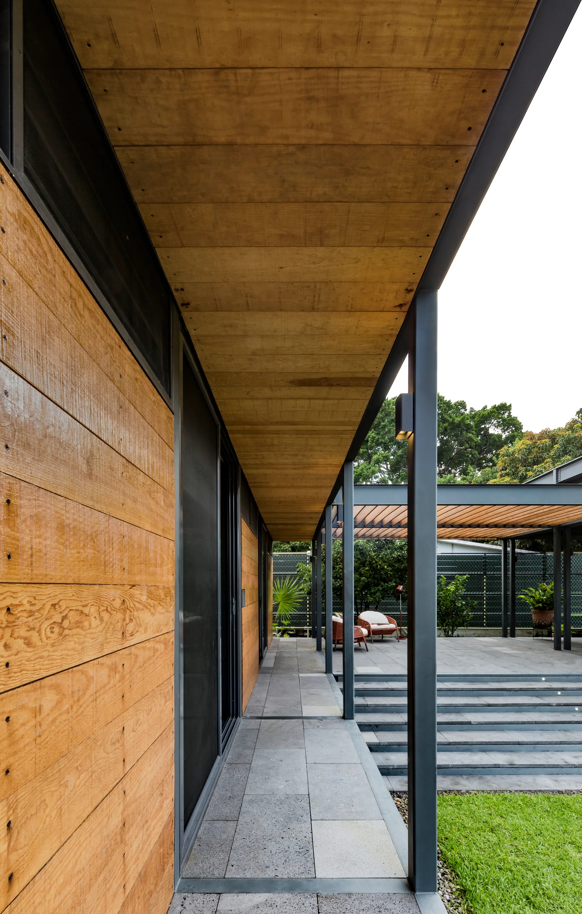 A modern covered outdoor patio featuring horizontal wood wall cladding, a wooden ceiling, black metal posts, and grey stone steps leading to a green lawn.