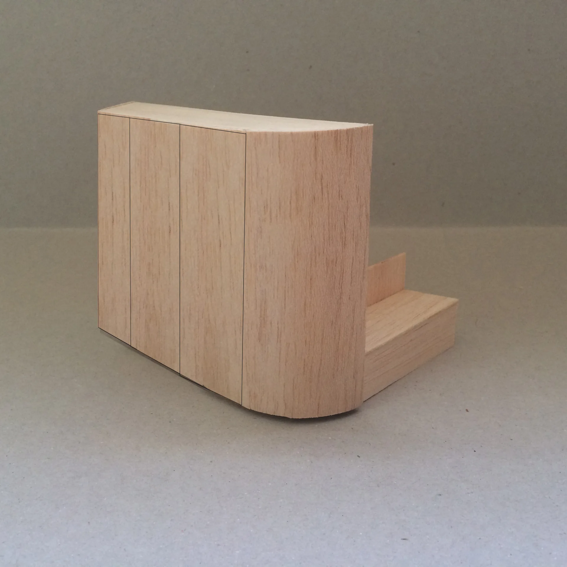 A wooden architectural scale model of a minimalist cabinet unit featuring a curved corner profile and vertical panel seams against a neutral background.