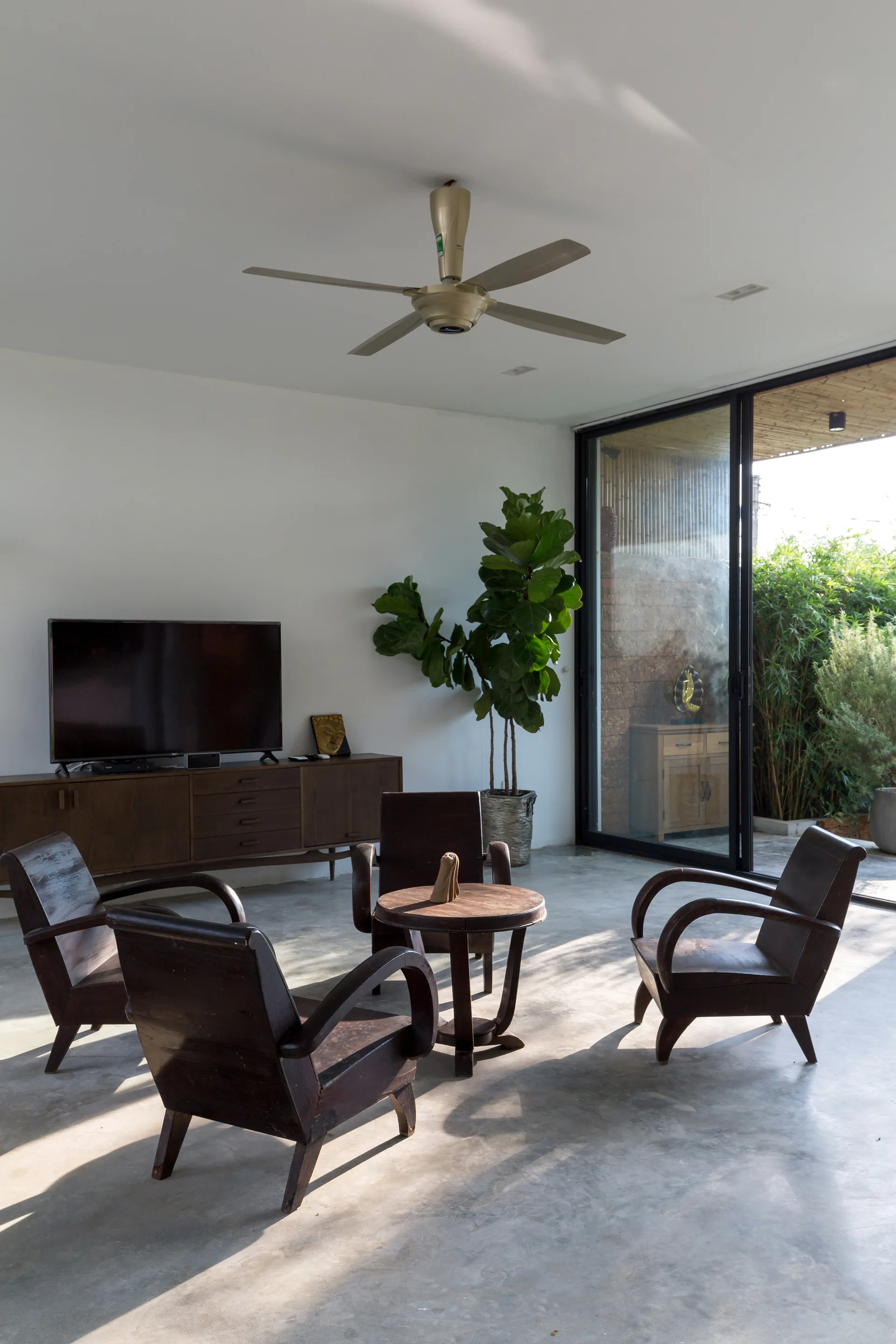 A bright living room features four dark wooden armchairs gathered around a small round table on a polished concrete floor next to sliding glass doors.