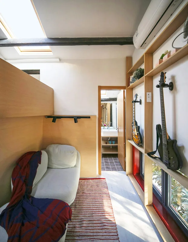 A compact room features a white sofa, a striped rug, and wall-mounted guitars on light wood built-in shelving beneath a skylight.