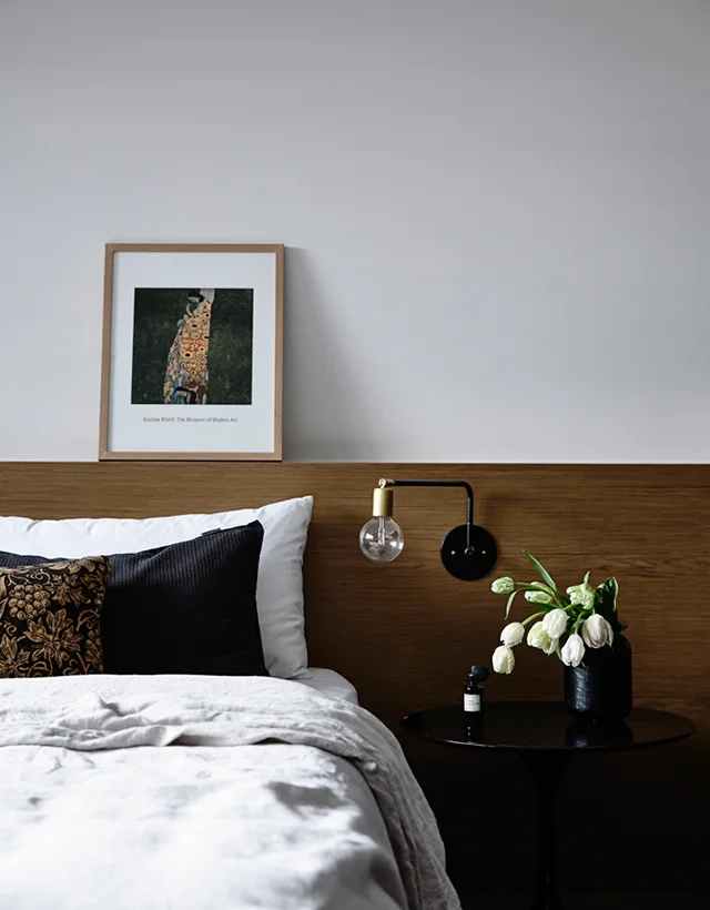 A modern minimalist bedroom featuring white linen bedding, a warm wood panel headboard, an integrated wall sconce, and a black round side table.