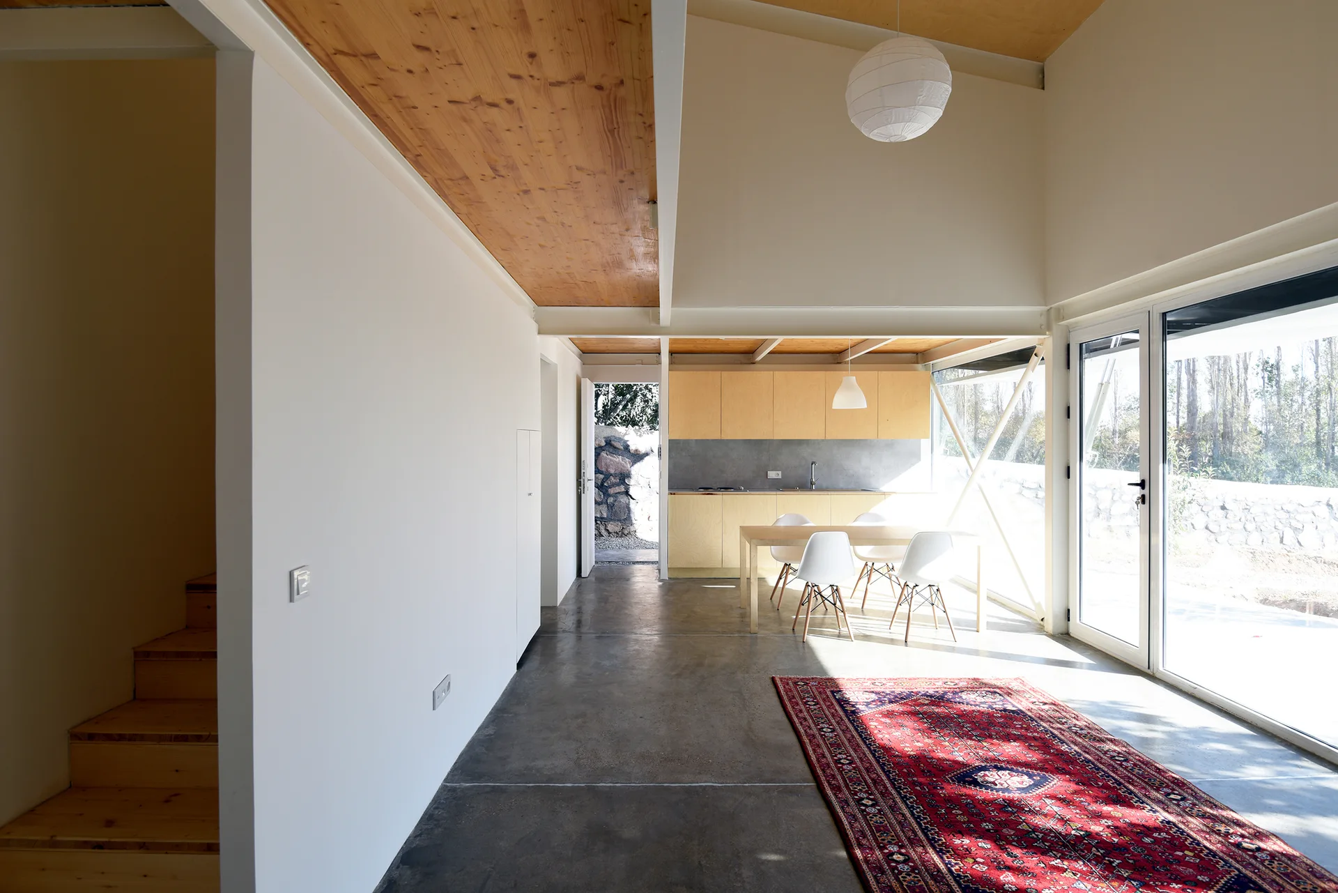 A modern open kitchen features light wood cabinets, a dining table with white chairs, and a red patterned rug on polished concrete floors.