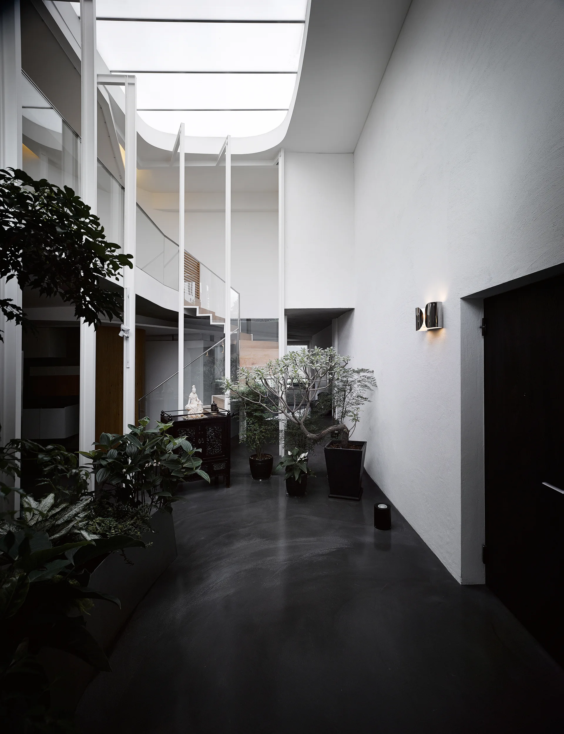 A high-ceilinged entryway features a large curved skylight, white walls, dark flooring, a glass-railed staircase, and potted green plants.