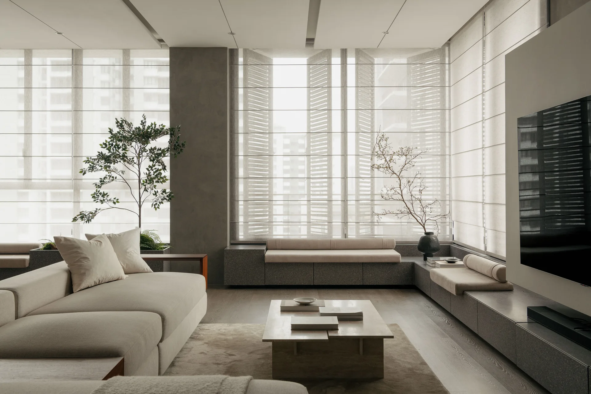 A minimalist living room with a beige sectional sofa, a low wooden coffee table, and built-in stone benches beneath windows with fabric shades.