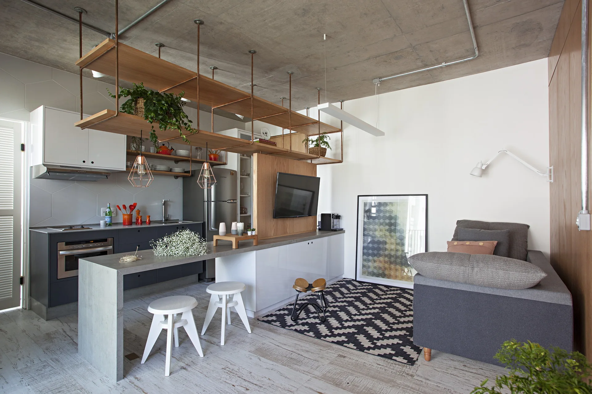 An open-plan kitchen and living space featuring a concrete ceiling, suspended wooden shelving, a grey island, and white stools.