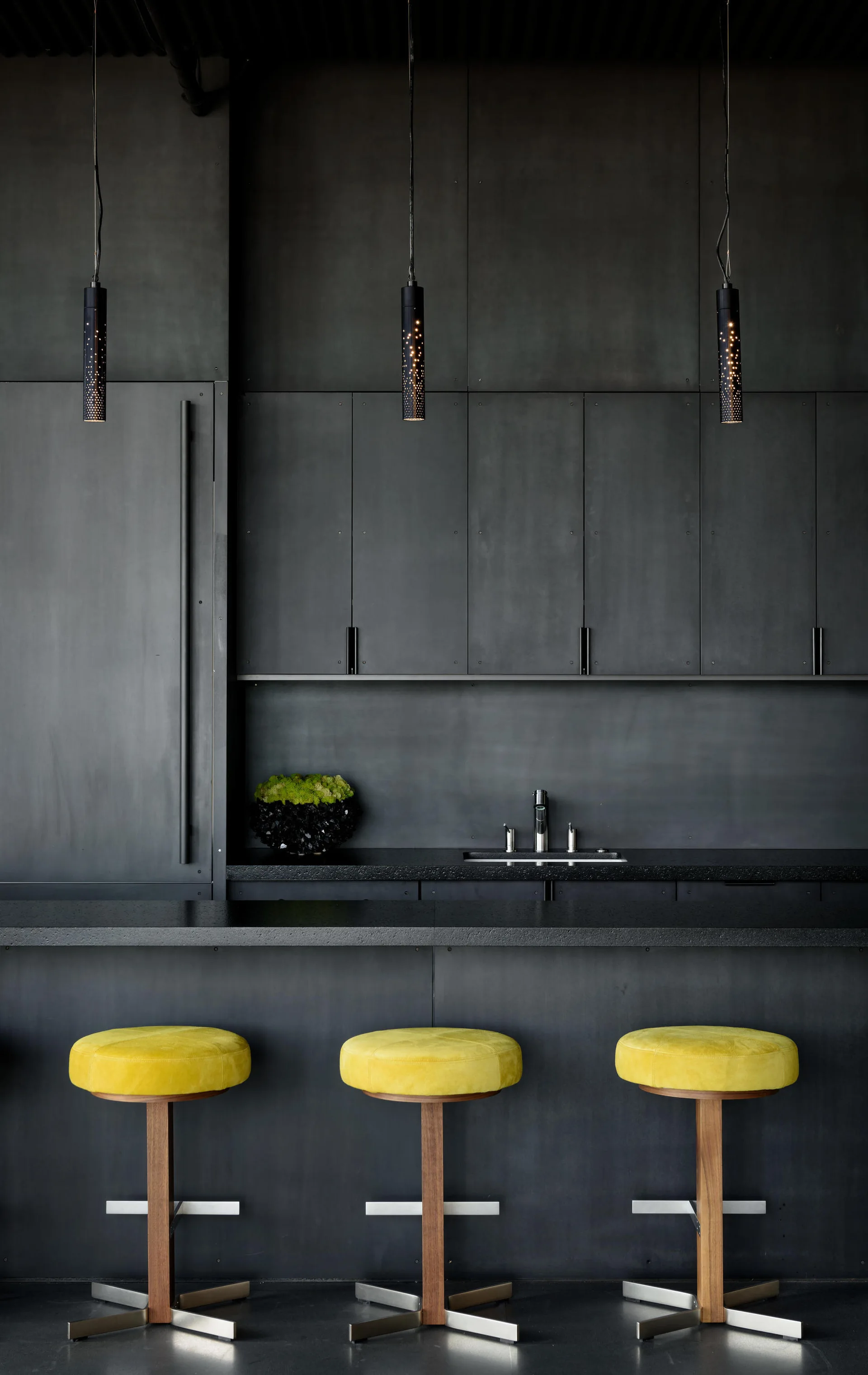 Three round yellow bar stools sit in front of a matte charcoal kitchen island counter and seamless black cabinets with cylindrical pendant lights.