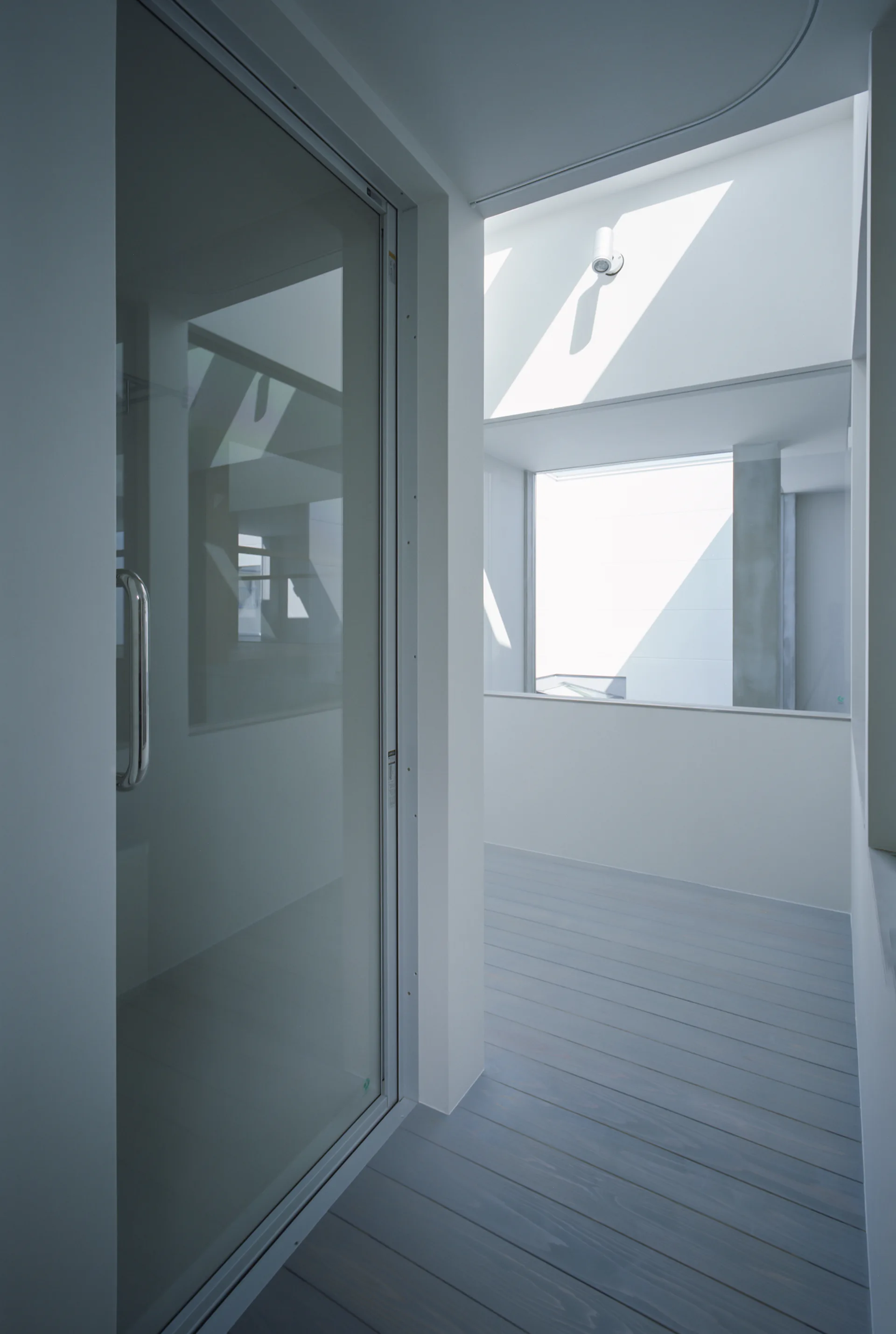 A minimalist indoor transition area with a sliding glass door opening to a balcony with grey wood deck flooring and bright white walls.