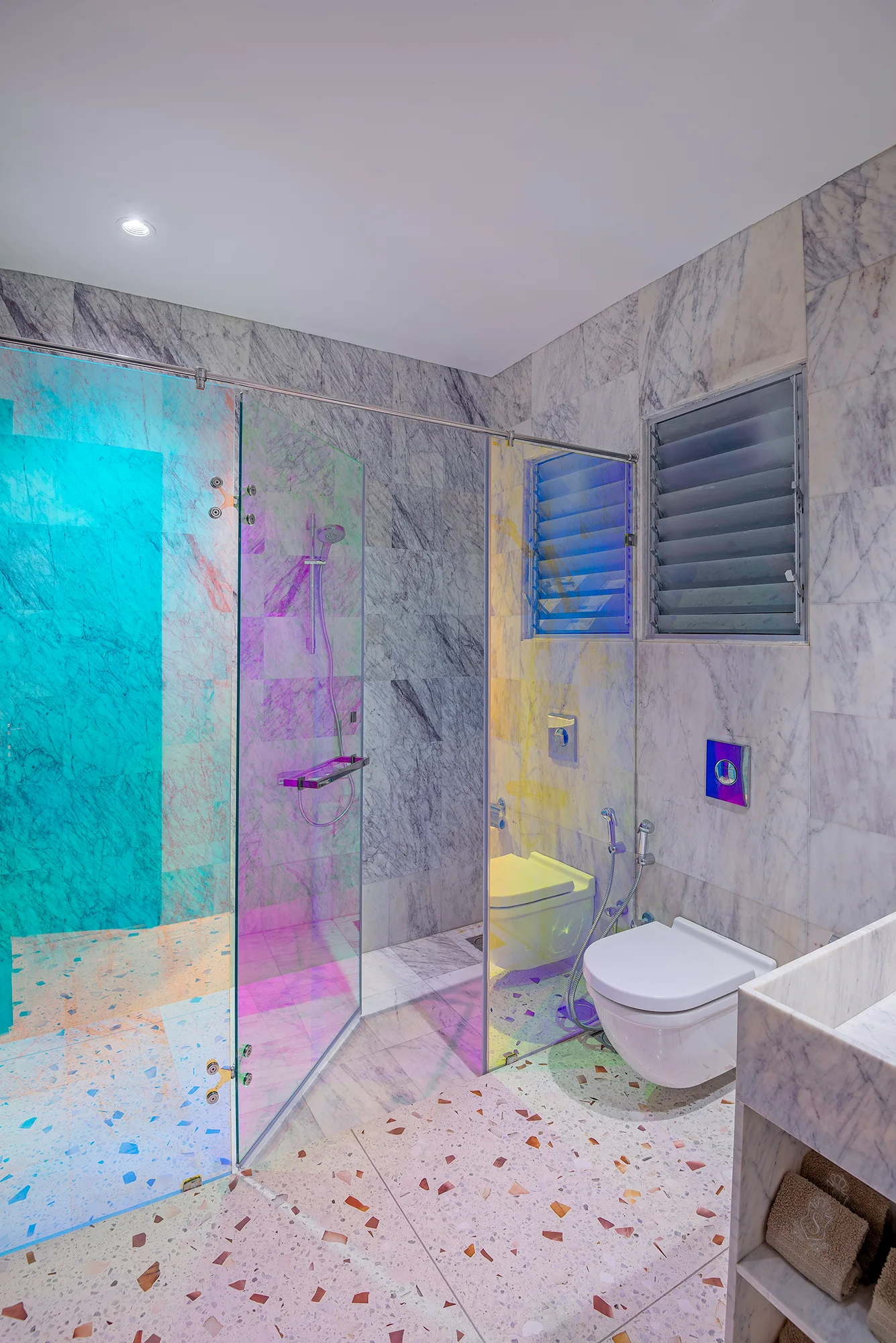 A modern bathroom featuring grey marble wall tiles, a speckled terrazzo floor, a wall-mounted toilet, and an iridescent glass shower partition.