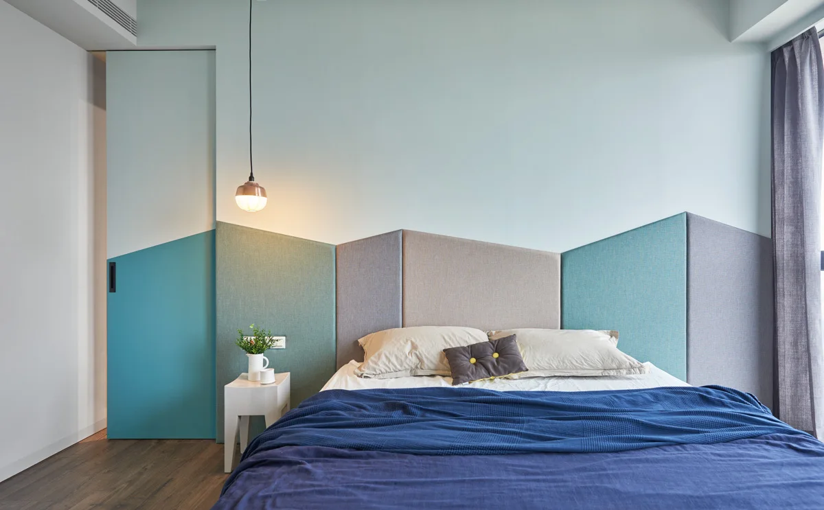 A modern bedroom features an asymmetrical upholstered headboard in blue and grey panels, a teal sliding door, and a low bed with blue linens.