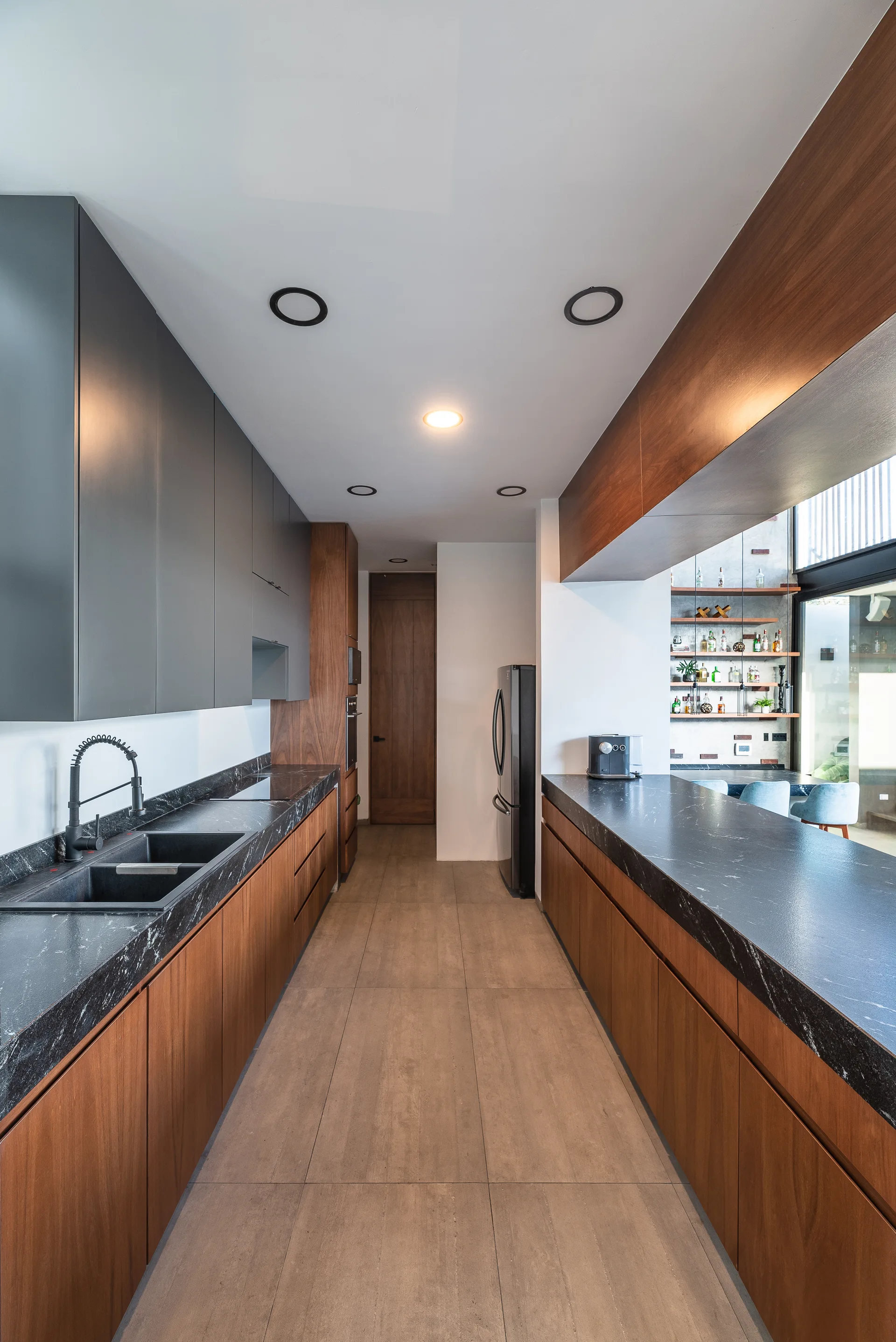 A modern galley kitchen featuring warm wood lower cabinets, matte gray upper cabinetry, dark veined stone countertops, and a black faucet.