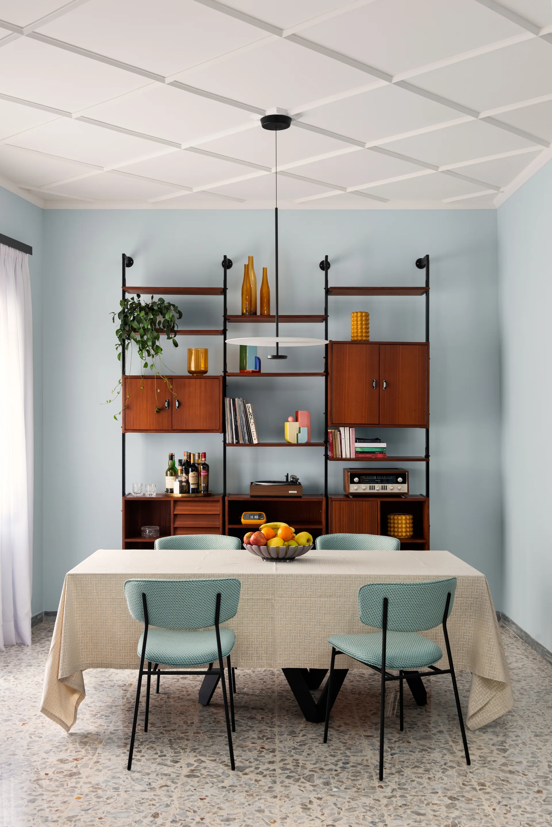 A dining room features a tablecloth-covered table with blue textured chairs beneath a white grid ceiling and a large modular wooden shelving unit.