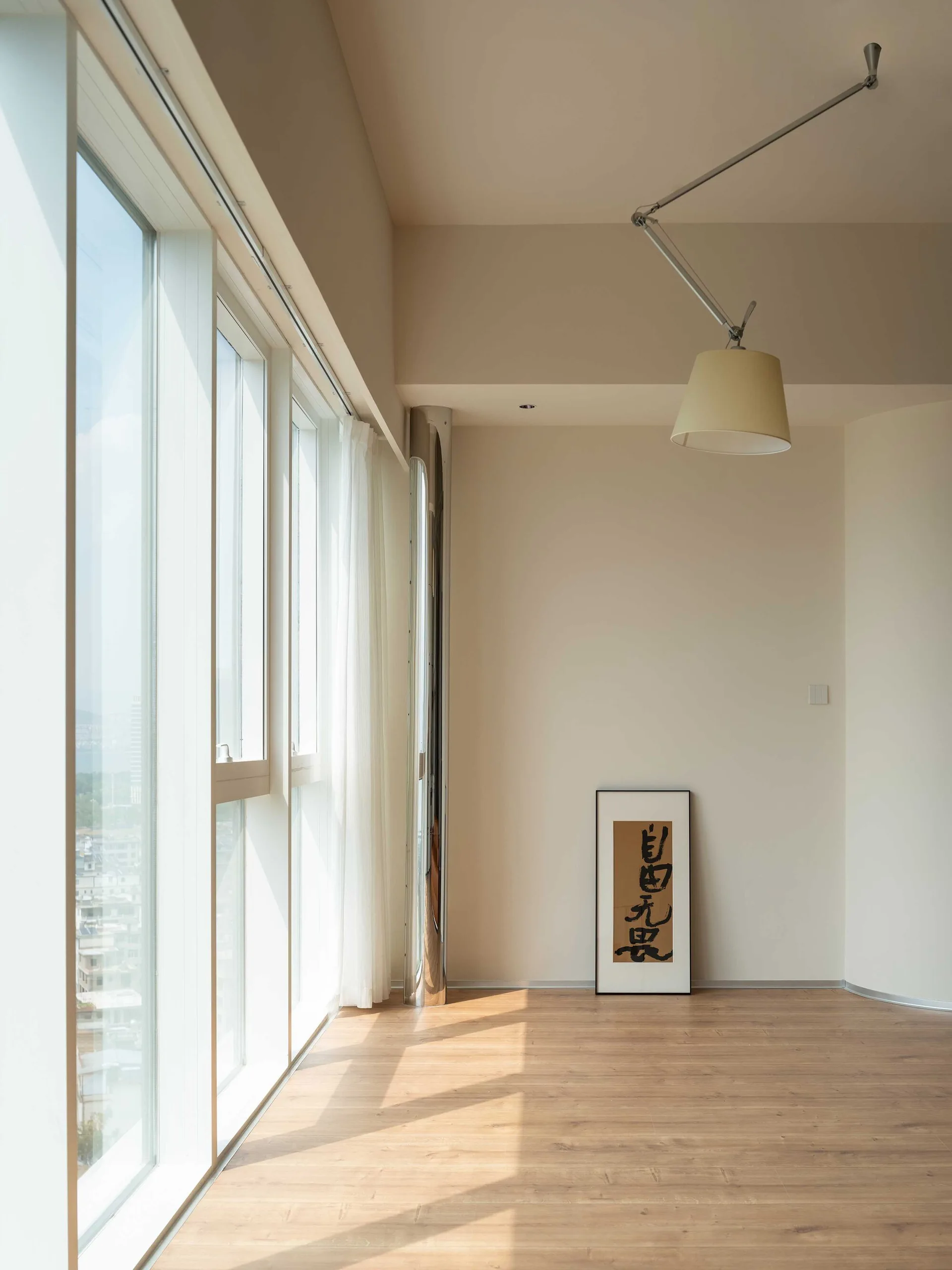 A minimalist room with light wood floors features sheers on large windows, a reflective chrome column, and an articulating arm ceiling light.