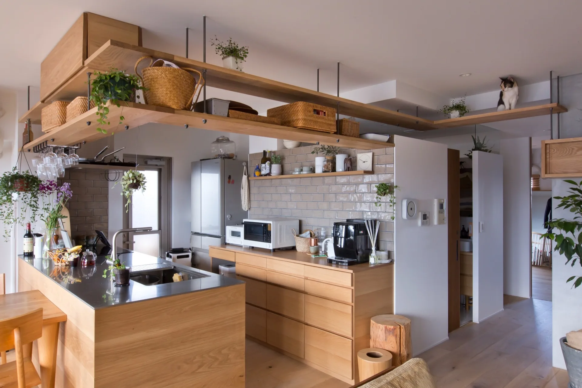 A minimalist kitchen features a light oak island with a stainless steel top, gray tiled walls, and a suspended wooden shelf holding woven baskets.