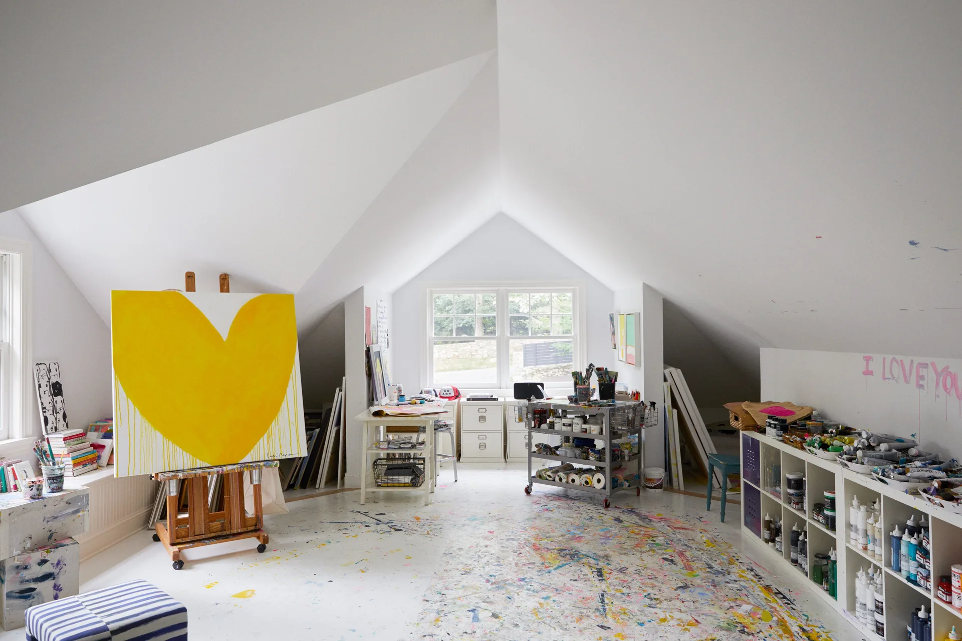 An attic art studio with sloped ceilings features a paint-splattered floor, a large yellow heart painting on an easel, and low white supply shelves.