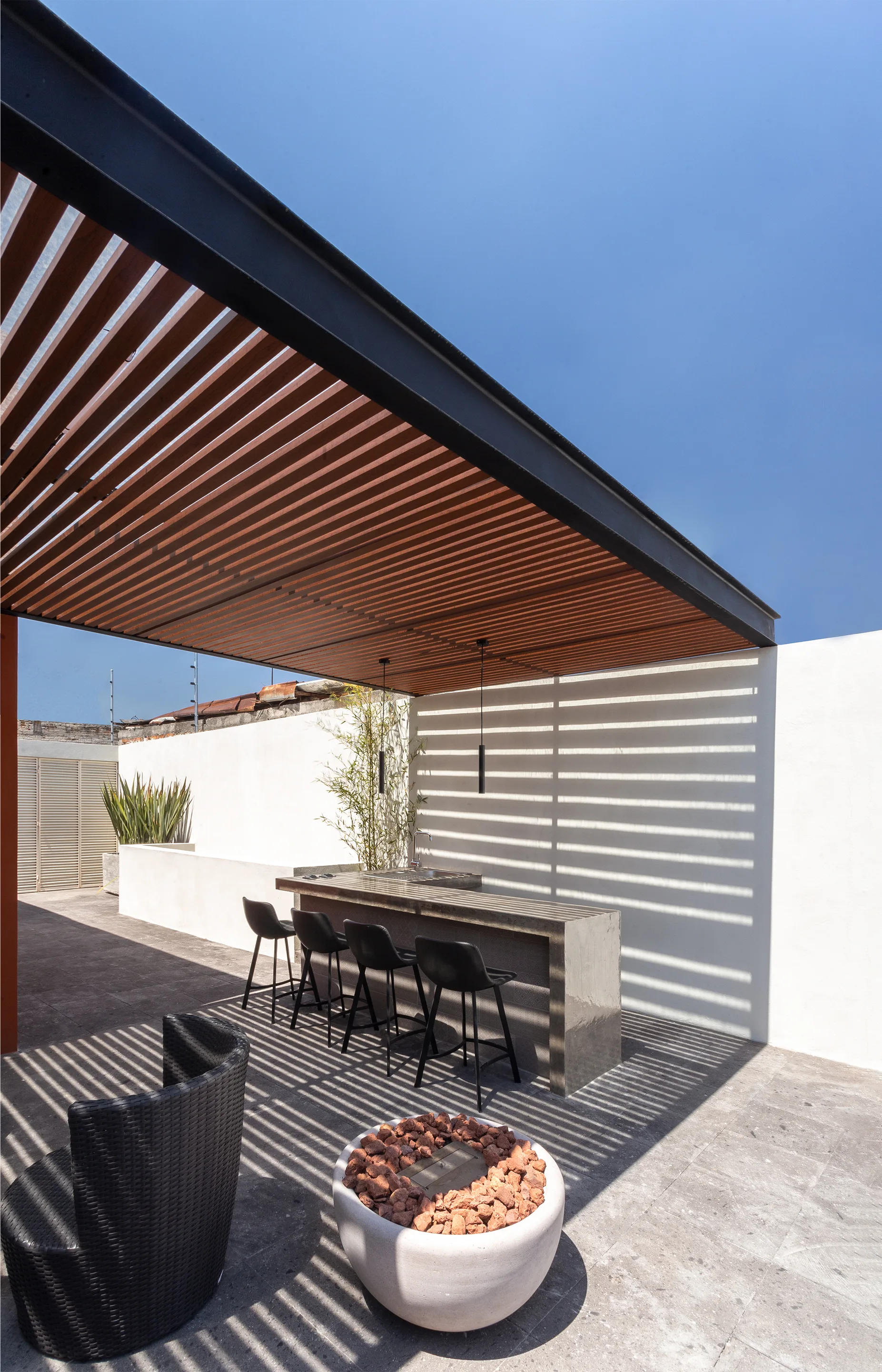 A modern patio features a concrete bar with black stools under a wood-slatted pergola, which casts linear shadows on a white wall.