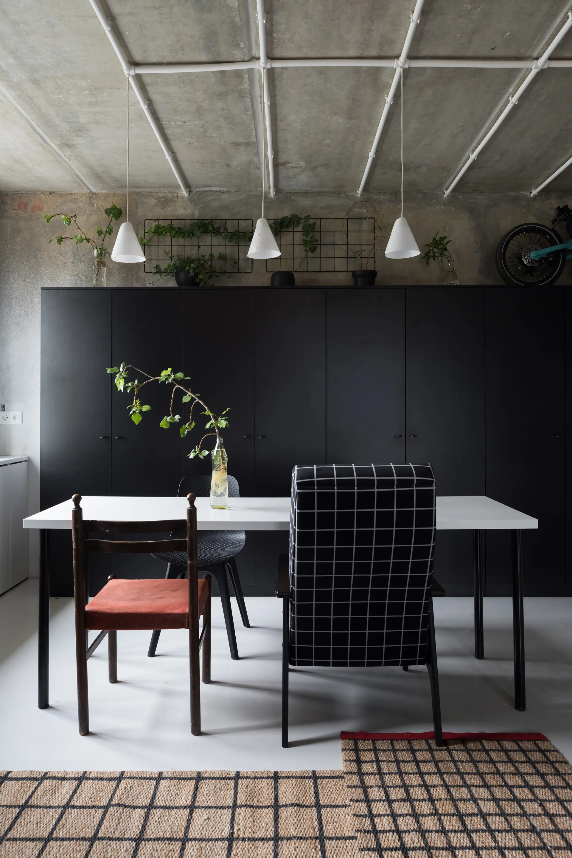 An industrial dining area features a white table, mismatched chairs, and three pendant lights against a wall of matte black cabinets.