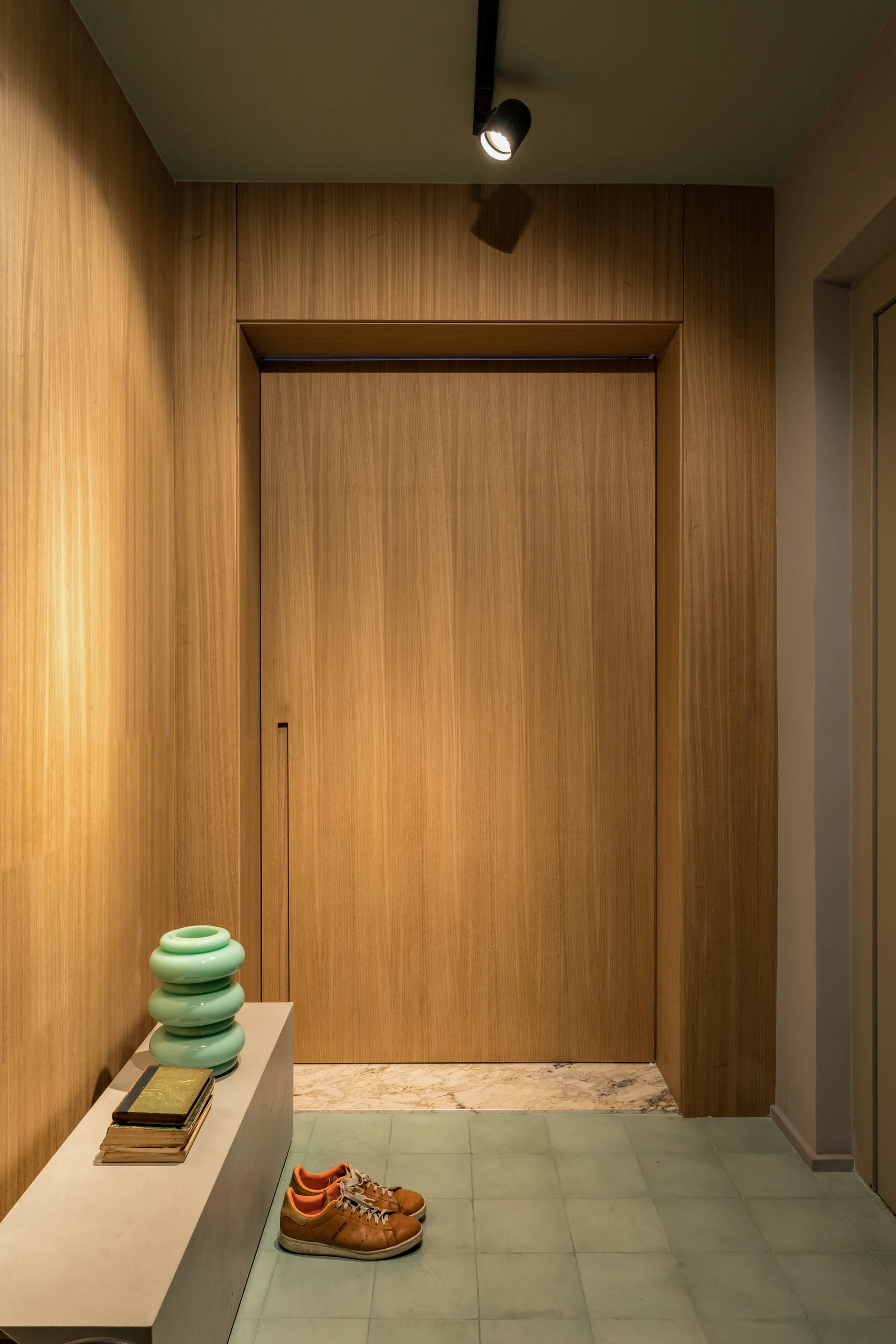 A minimalist entryway features a flush wood-paneled door, sage green square floor tiles, and a low concrete bench with a green glass vase.