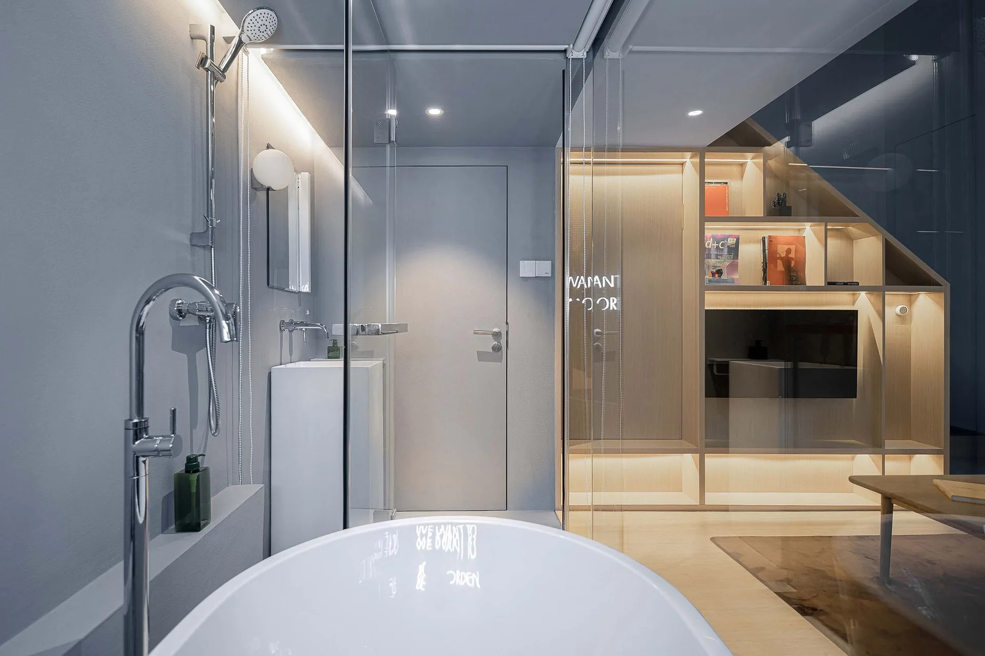 A minimalist bathroom featuring a white freestanding bathtub, a glass partition, and a grey flush door next to built-in illuminated wood shelving.