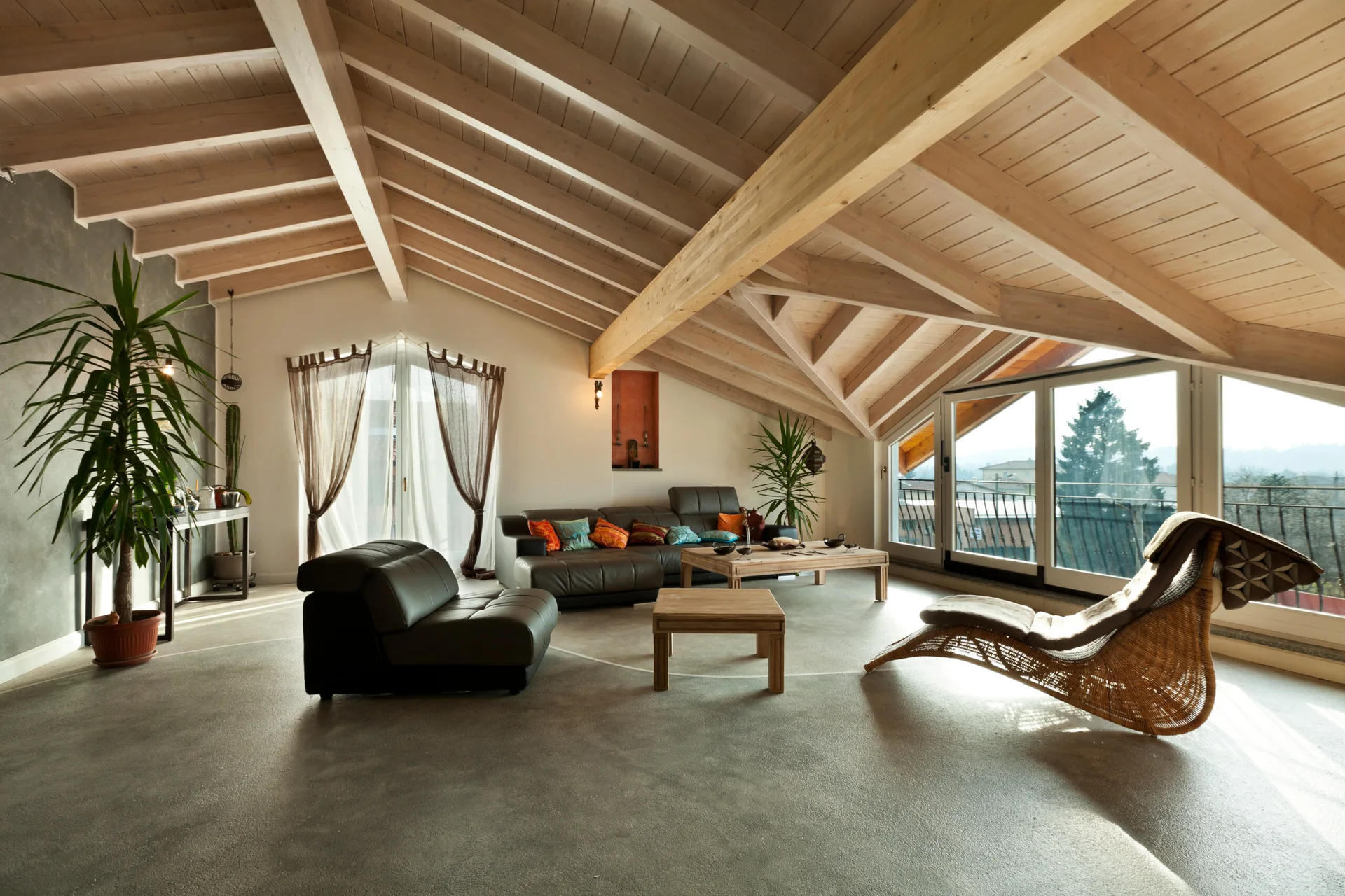 A spacious attic living room features a pitched ceiling with exposed wood beams, dark leather sofas, a wicker chaise lounge, and gray flooring.