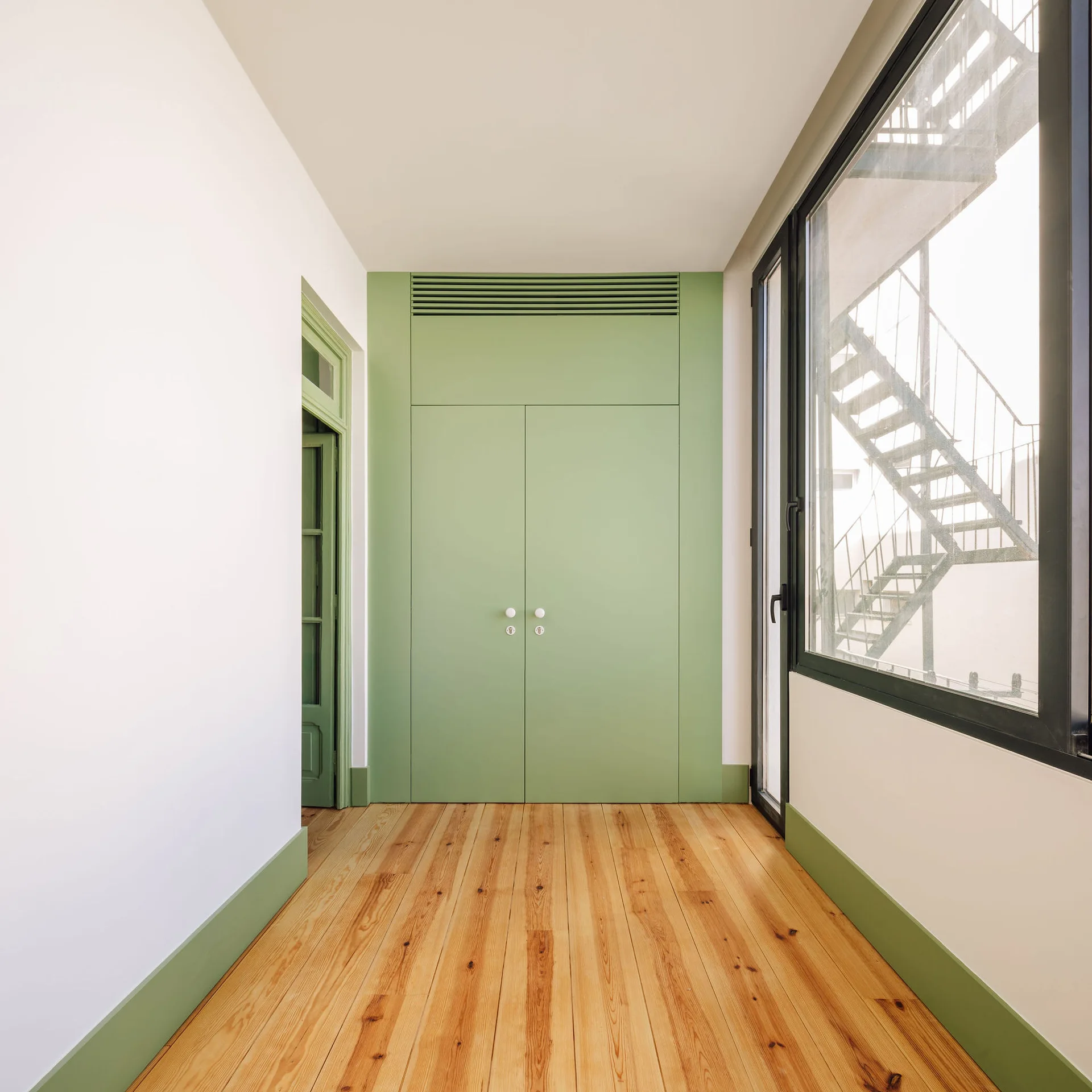 A minimalist hallway with light wood floors features a tall sage green built-in closet, matching green baseboards, and a large side window.