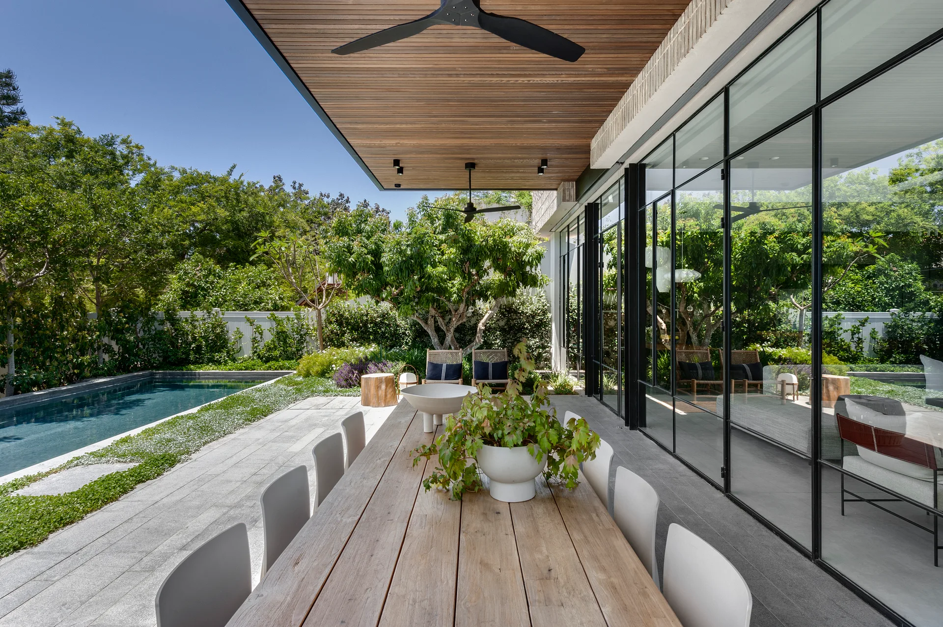 A covered patio features a long wood dining table, a slatted timber ceiling with a black fan, and large black-framed windows next to a swimming pool.