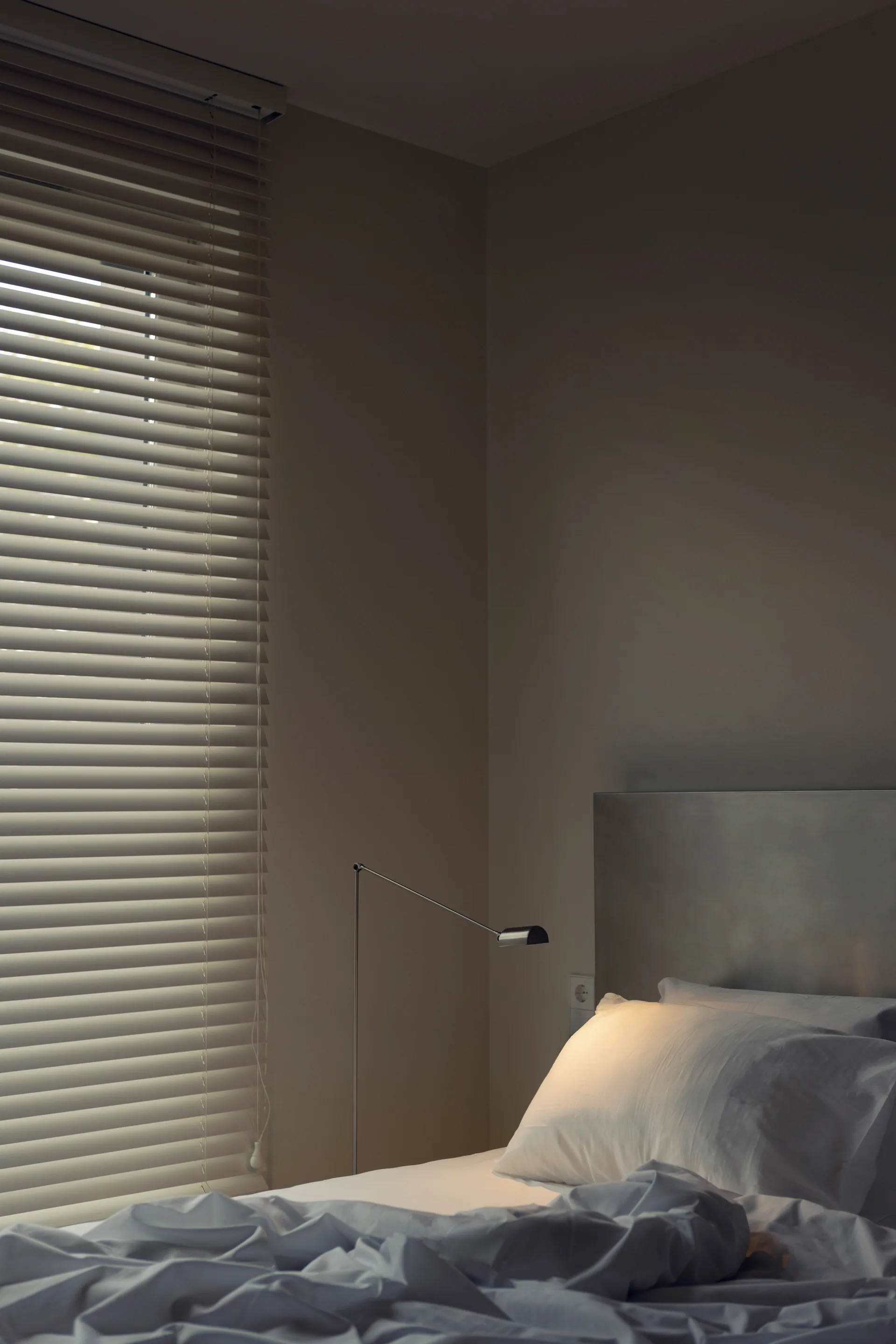 A minimalist bedroom features a bed with white linens, a brushed metal headboard, a slim adjustable reading lamp, and window blinds.