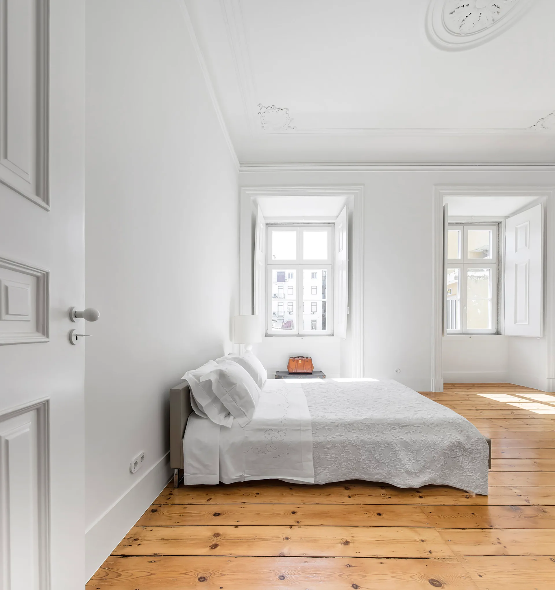 A bright minimalist bedroom features a low bed with white bedding, natural wide-plank wood floors, and traditional white window shutters.
