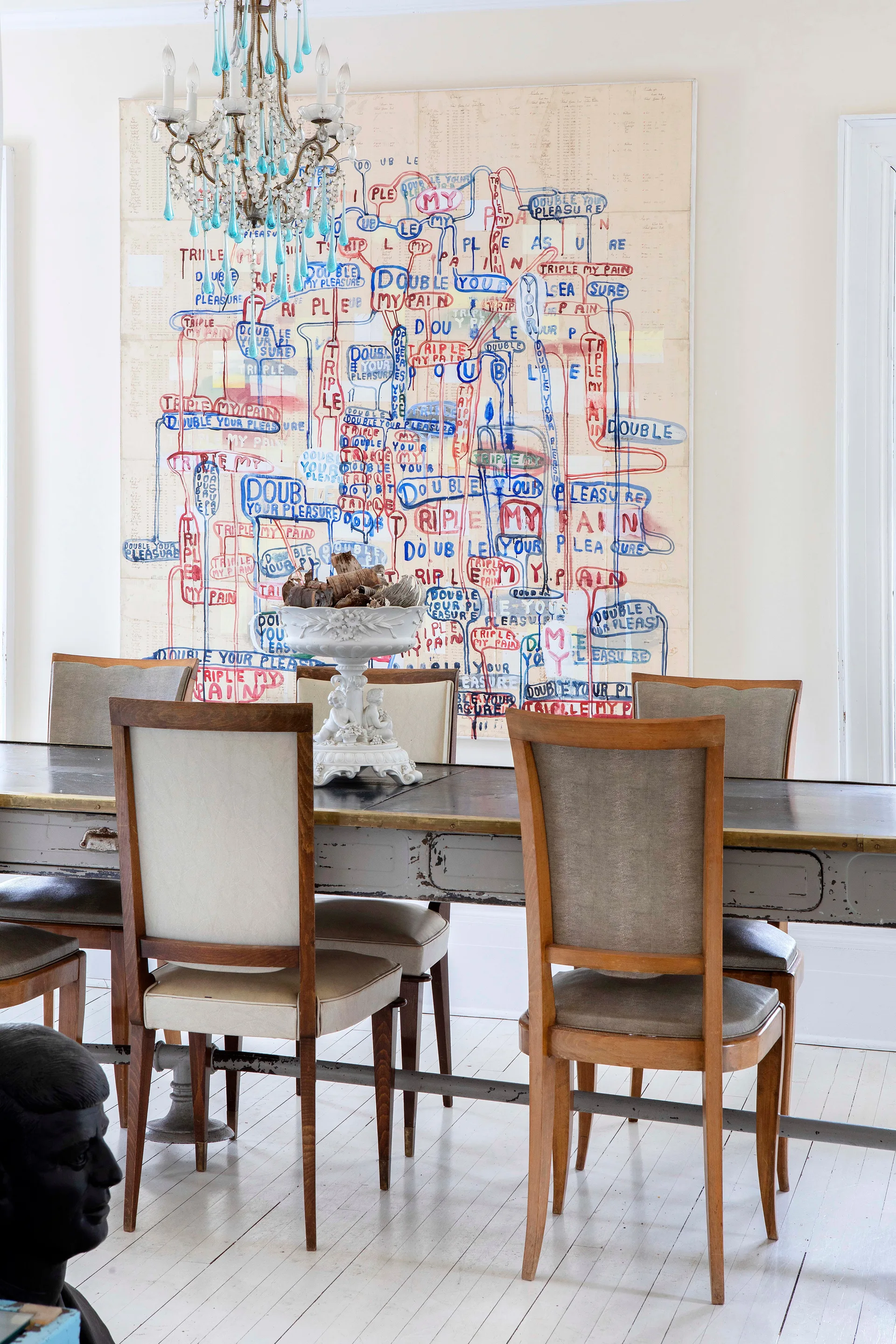 Wooden chairs surround a distressed dining table beneath a blue crystal chandelier and a large canvas printed with colorful typography.
