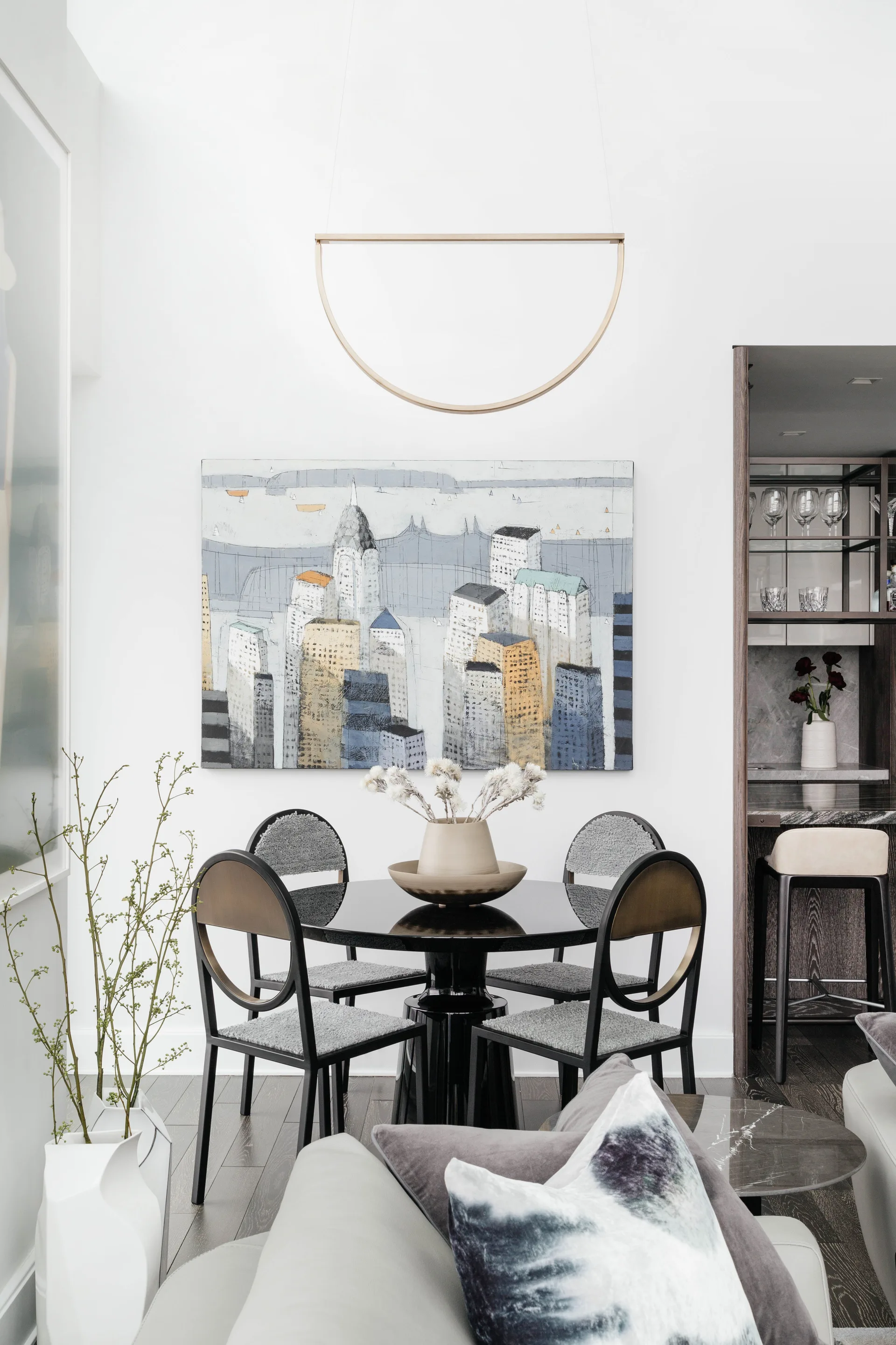 A high-gloss black round dining table is surrounded by four grey upholstered chairs under a city skyline painting and a gold half-moon pendant light.